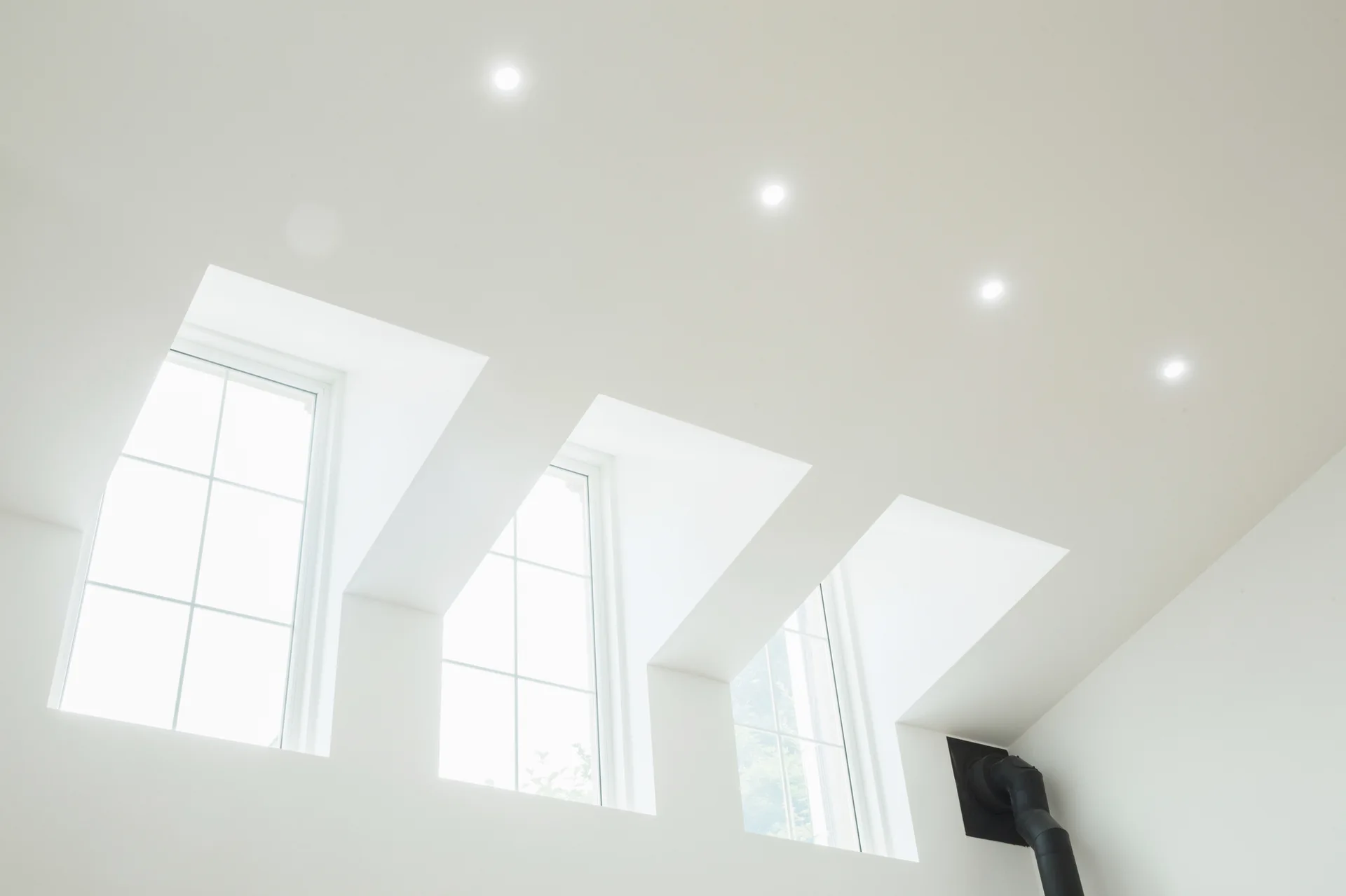 Three deeply recessed white windows line a sloped white ceiling with small recessed spotlights above.