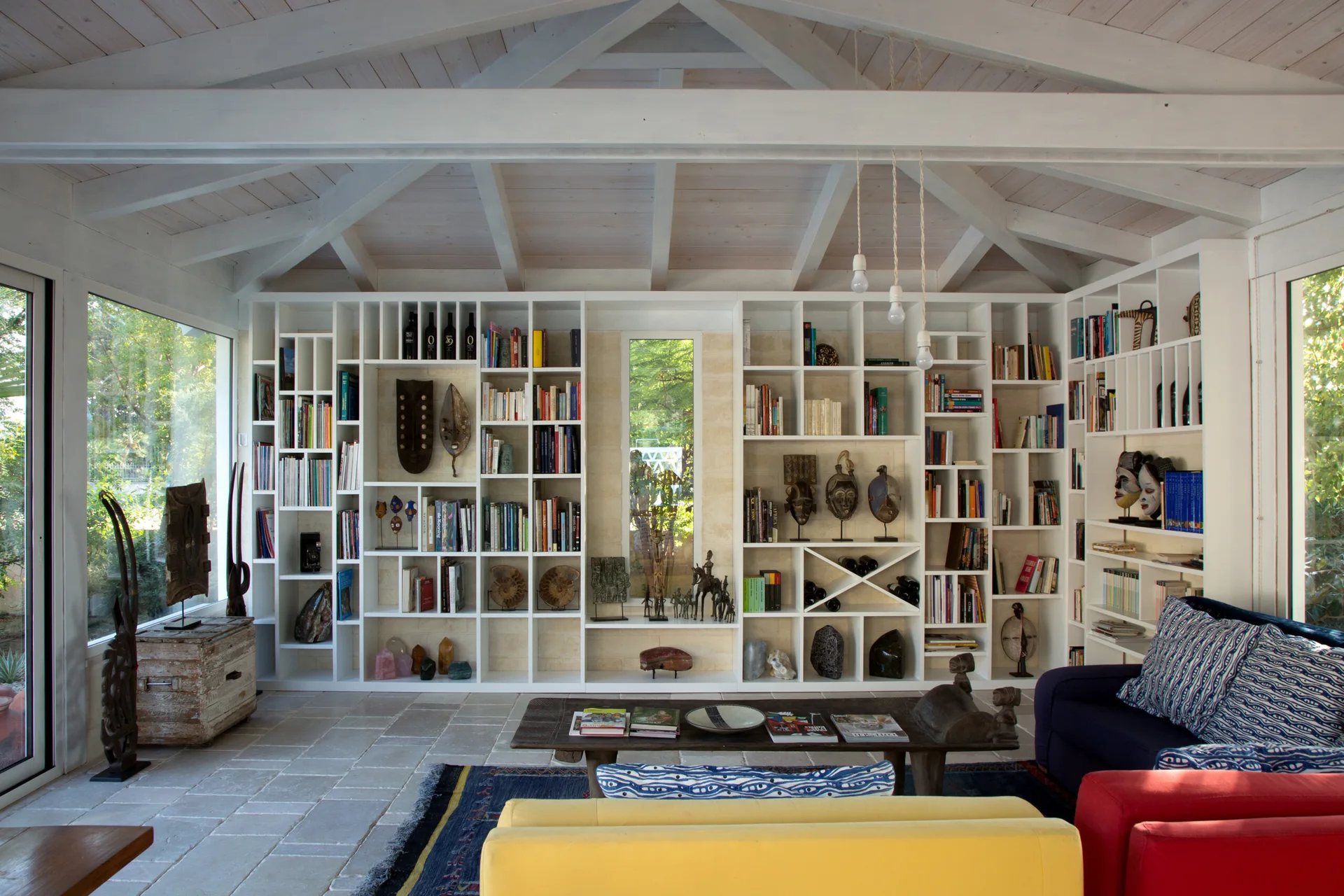 A bright living room features a white vaulted ceiling with exposed beams, a wall-to-wall display bookshelf, and colorful sofas on a gray stone floor.