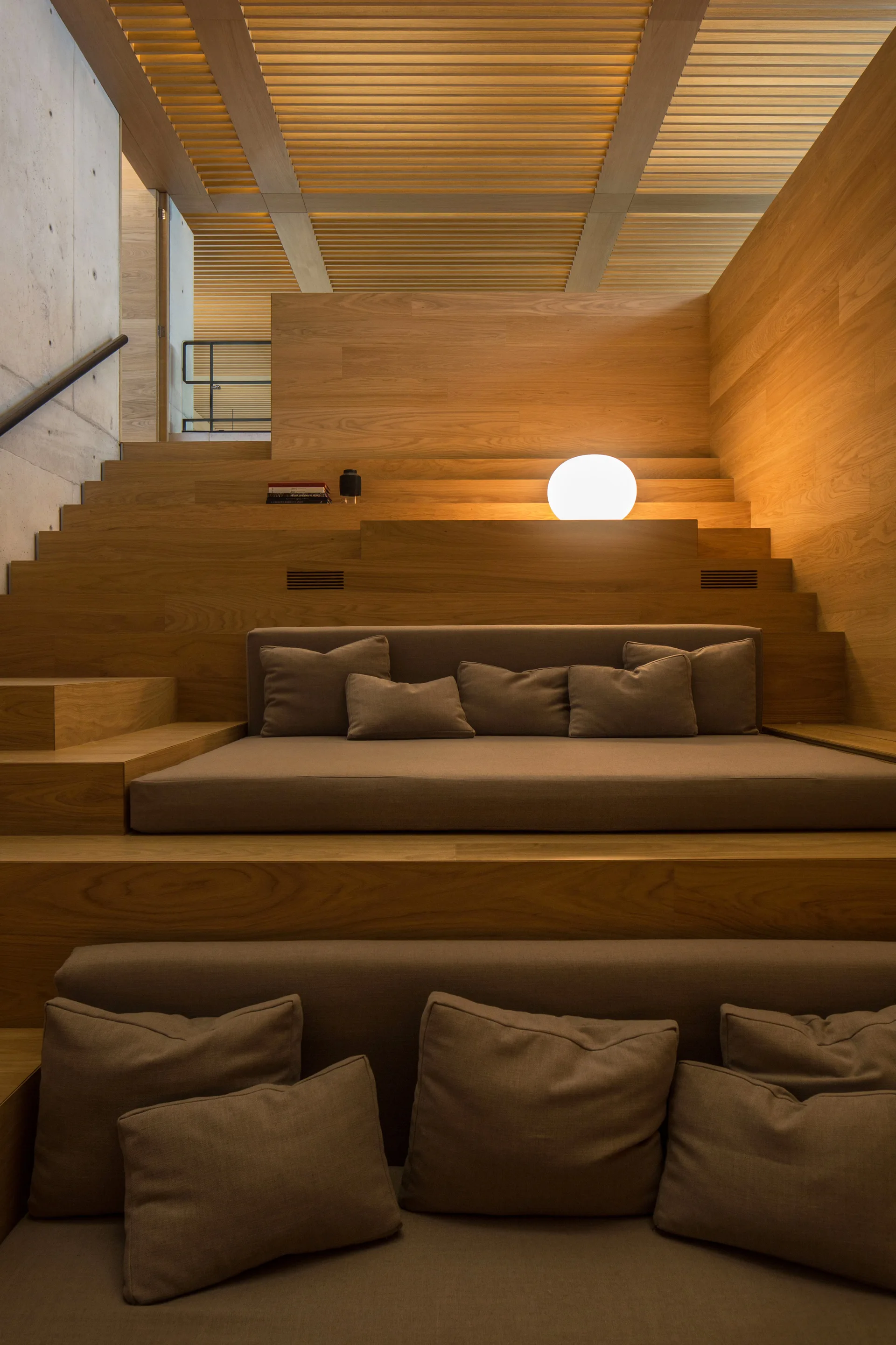 Built-in tiered wooden steps function as a lounge area with grey cushions, a glowing spherical lamp, and a slatted timber ceiling.