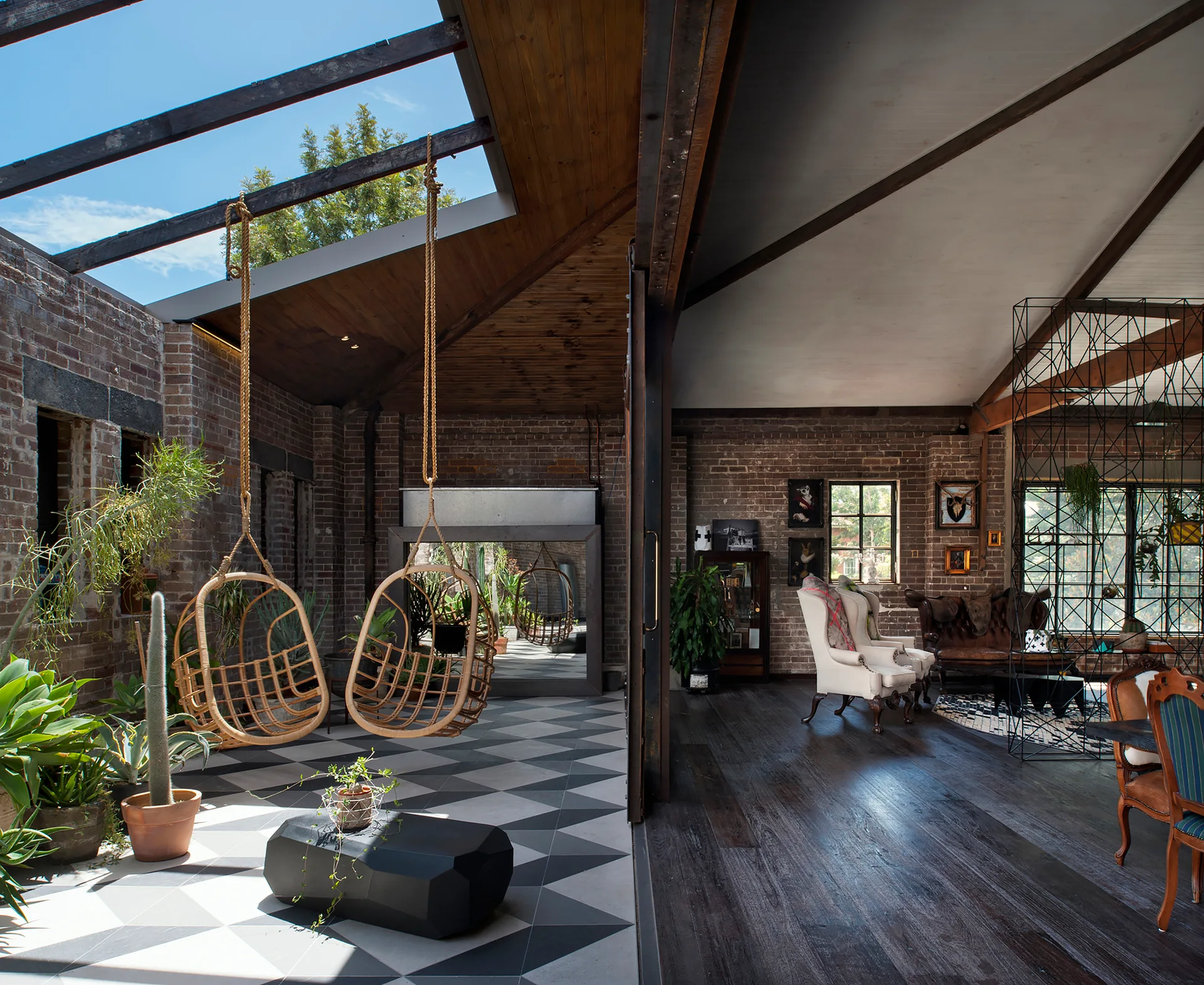 An industrial loft sunroom features hanging rattan chairs and plants on geometric floor tiles next to a wood-floored living area.