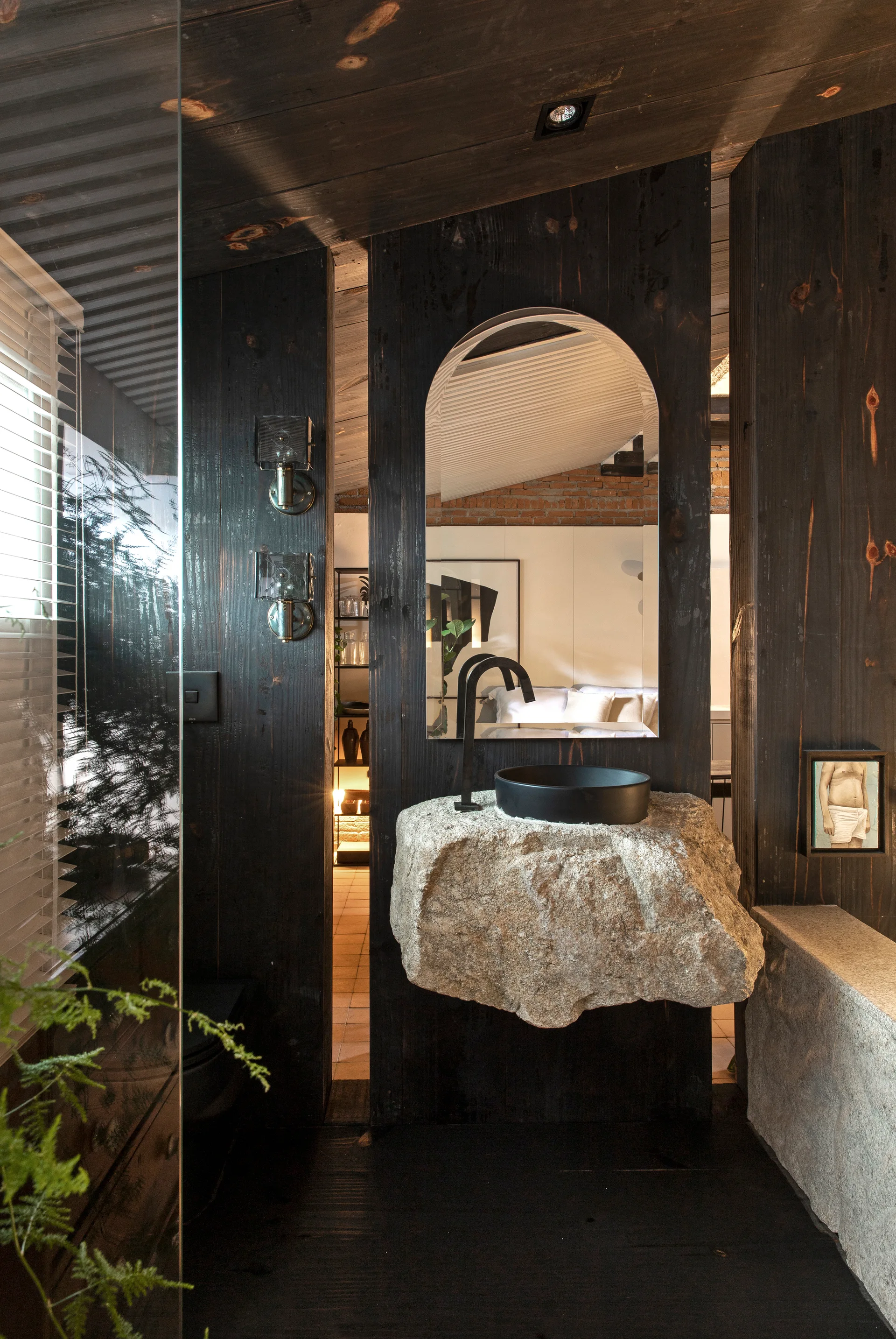 A modern rustic bathroom features a rough-cut stone vanity supporting a black vessel sink against dark vertical wood paneling.