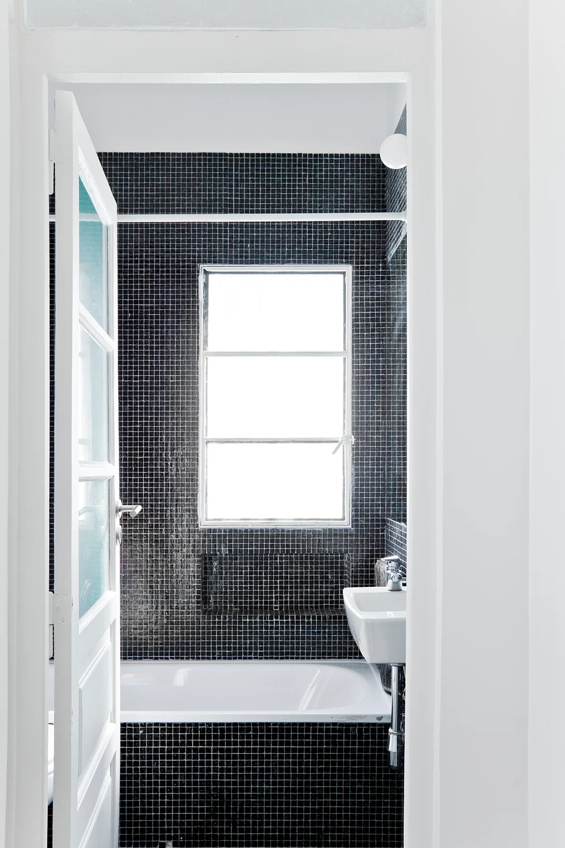 A bathroom viewed through a white doorway features black square mosaic tile walls, a central window, and a wall-mounted white ceramic sink.