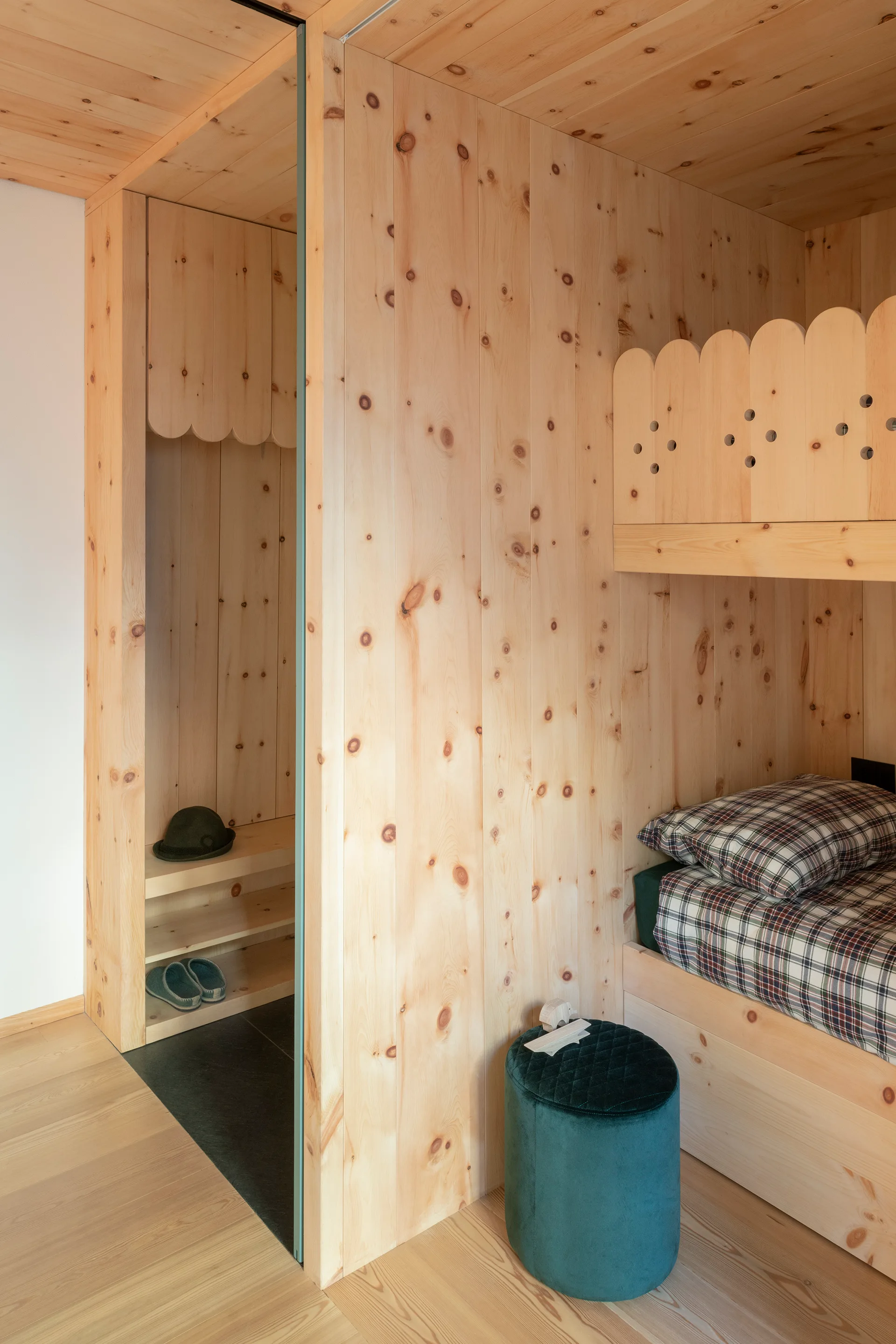 A compact bedroom lined in knotty pine wood paneling features a built-in bunk bed with a scalloped railing next to a green velvet stool.