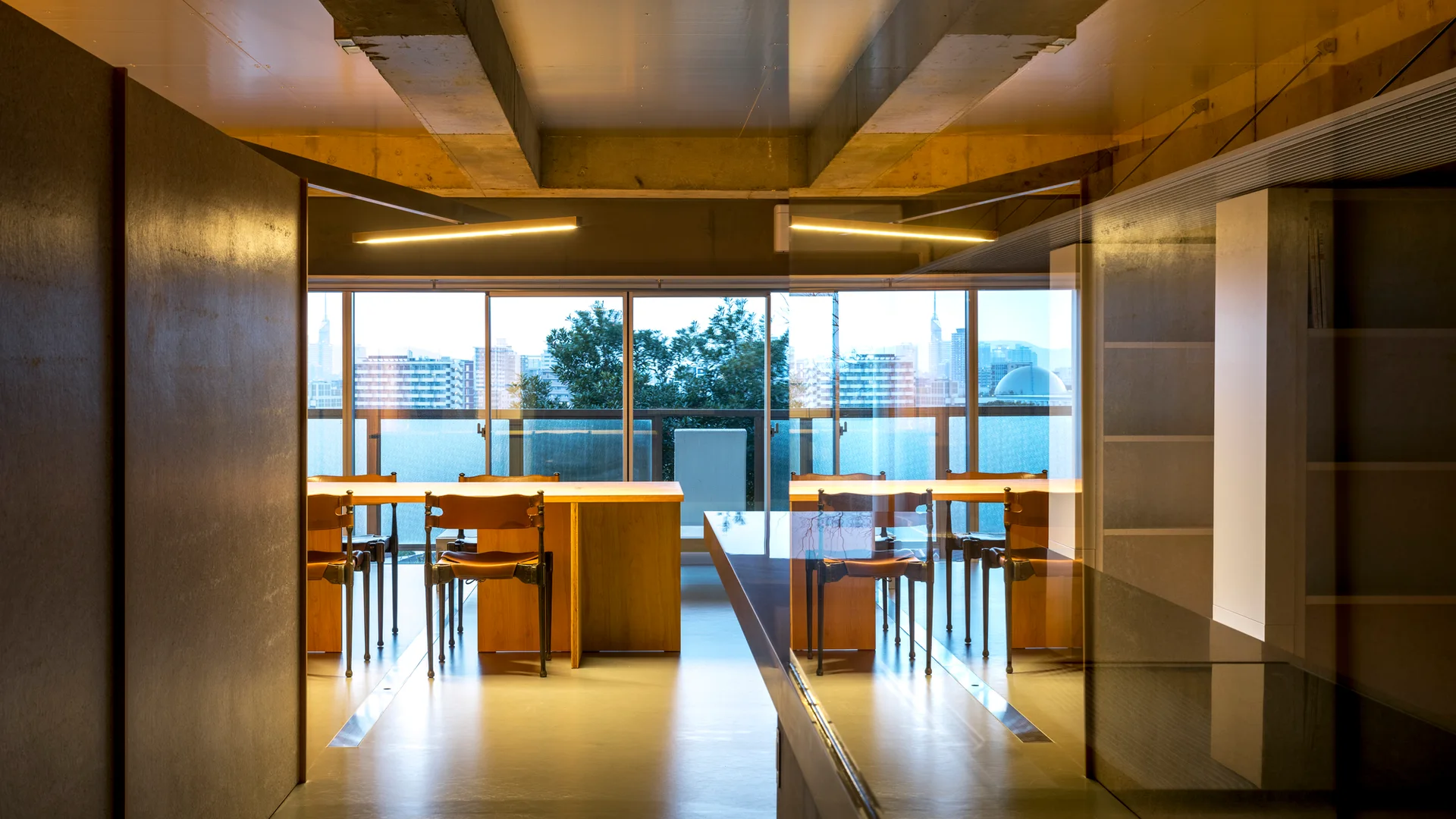 Wooden dining tables and leather chairs sit beneath an exposed concrete ceiling with linear pendant lights and large windows overlooking a city.