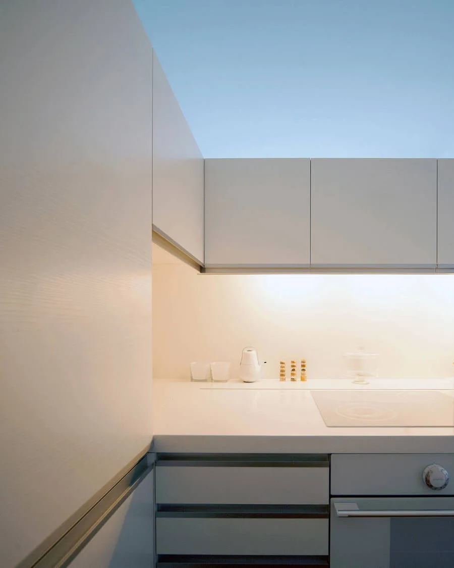 Minimalist white kitchen features flat-front upper and lower cabinets, a seamless countertop with an integrated cooktop, and warm under-cabinet lighting.