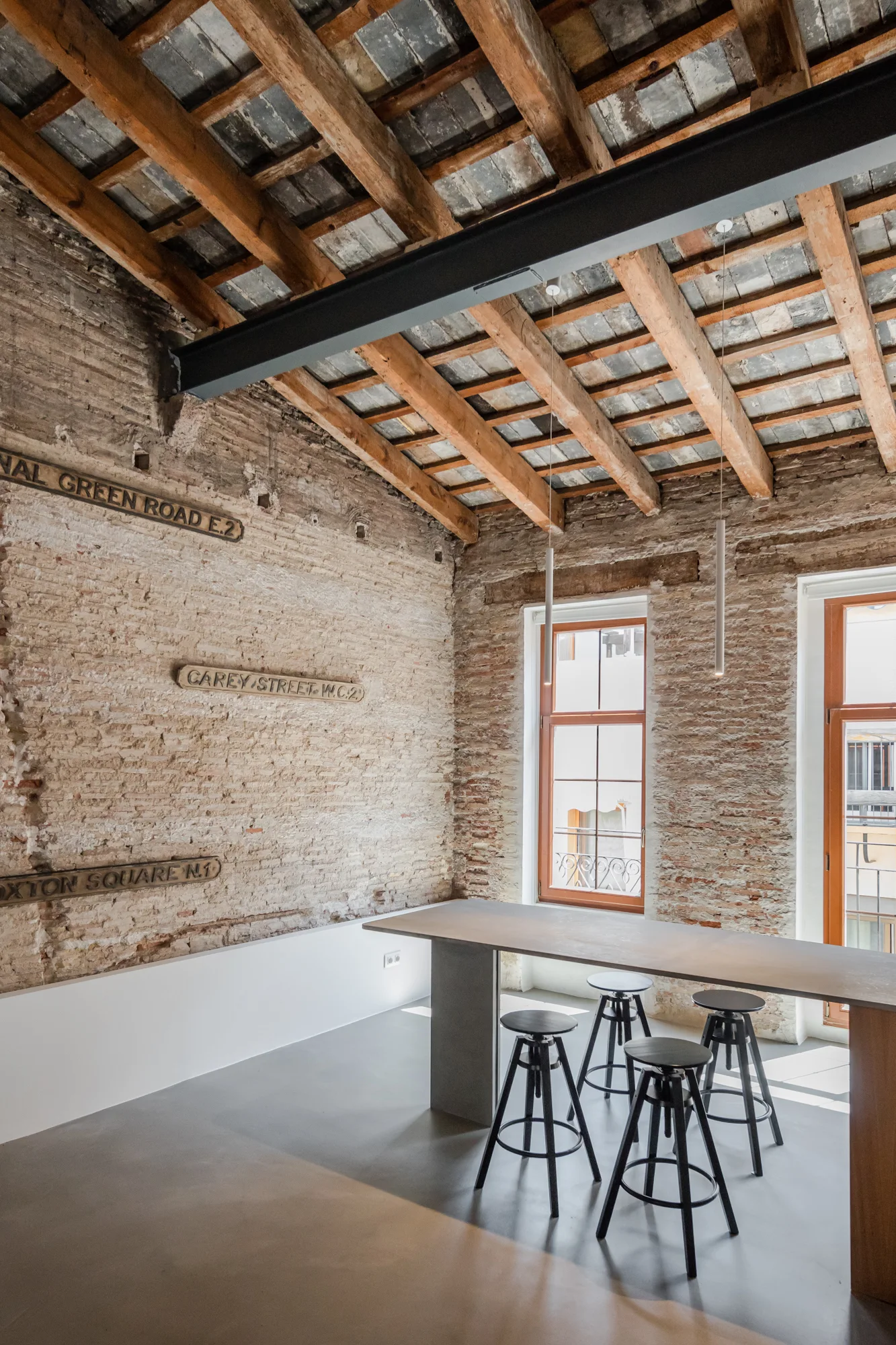 An industrial loft dining area with exposed brick walls, a wooden beamed ceiling, a grey concrete table, and four adjustable black metal stools.