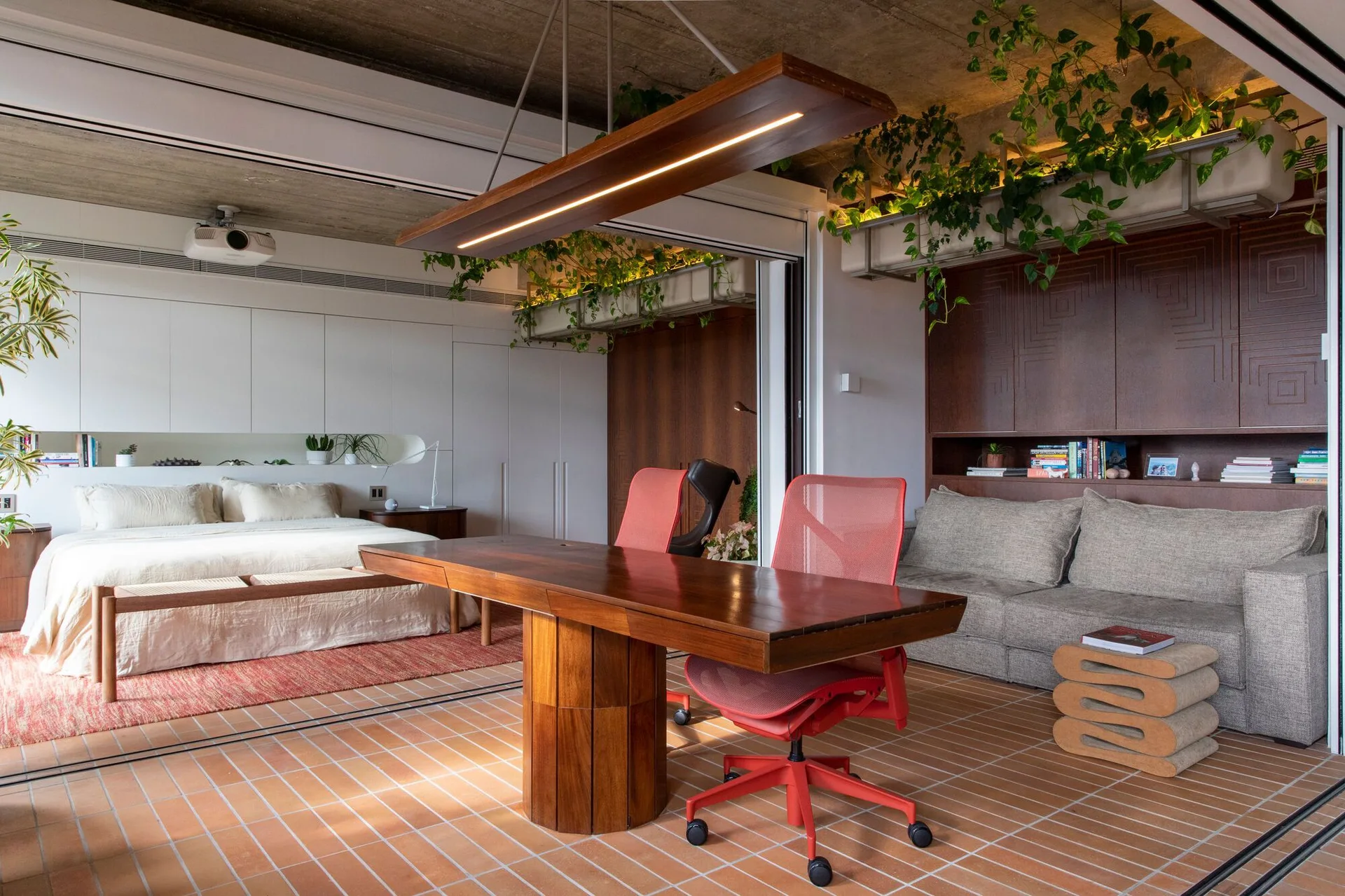 A multi-functional studio apartment featuring a long wood desk with red office chairs, terracotta tiled flooring, and a bedroom in the background.