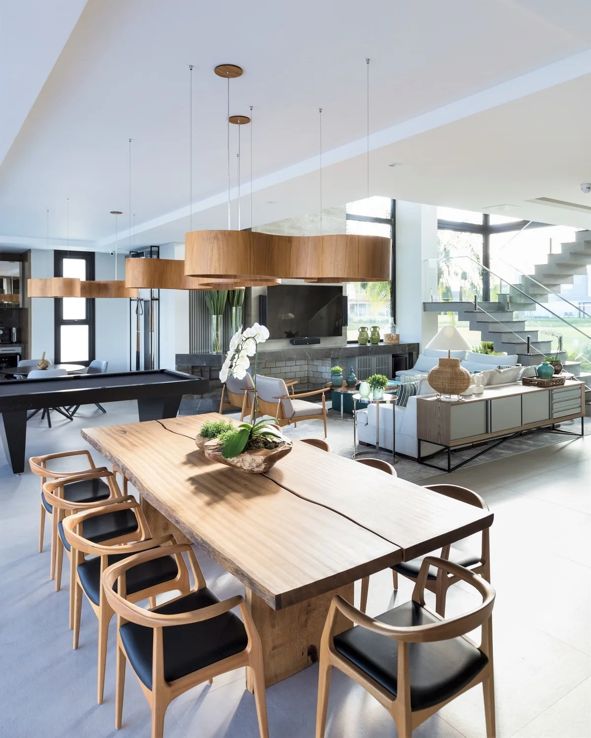 An open-concept dining area features a large live-edge wood table, modern wooden chairs with black seats, and sculptural wave-shaped wood pendant lights.