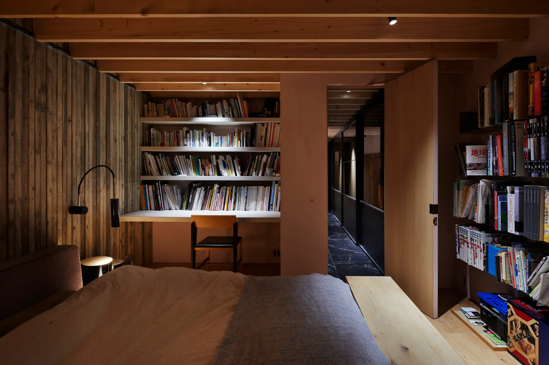 A bedroom with exposed wooden ceiling beams features a built-in desk and bookshelves under warm lighting, with a bed in the foreground.