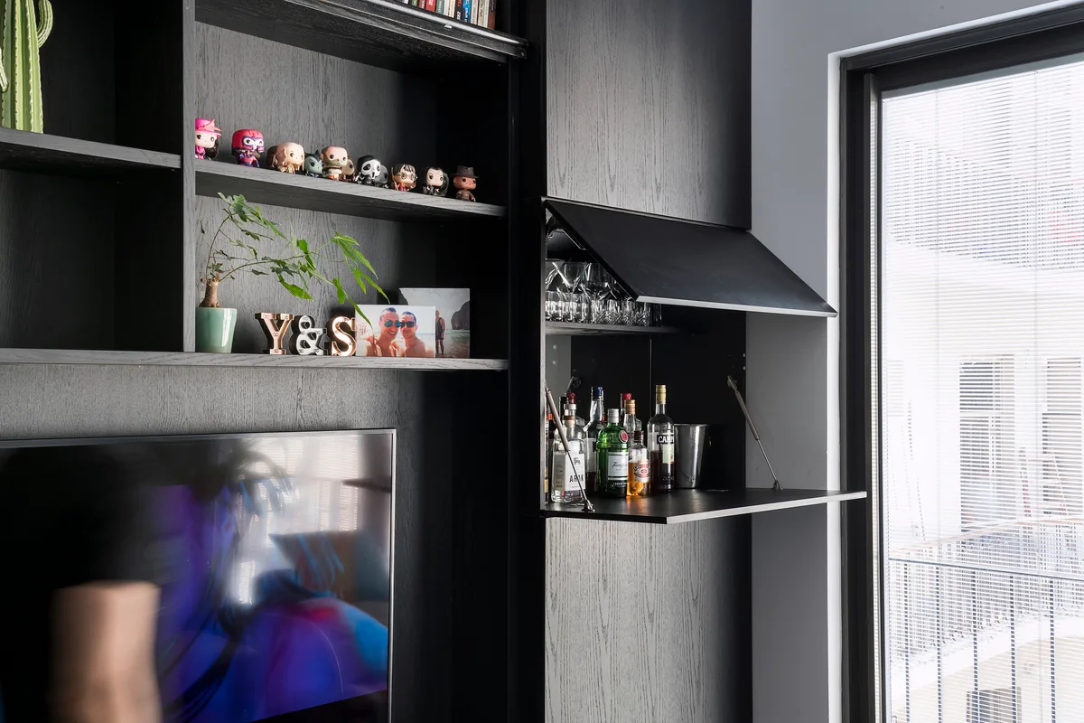 A matte black wood built-in shelving unit with decorative items next to an open dropdown cabinet used as a home liquor bar.