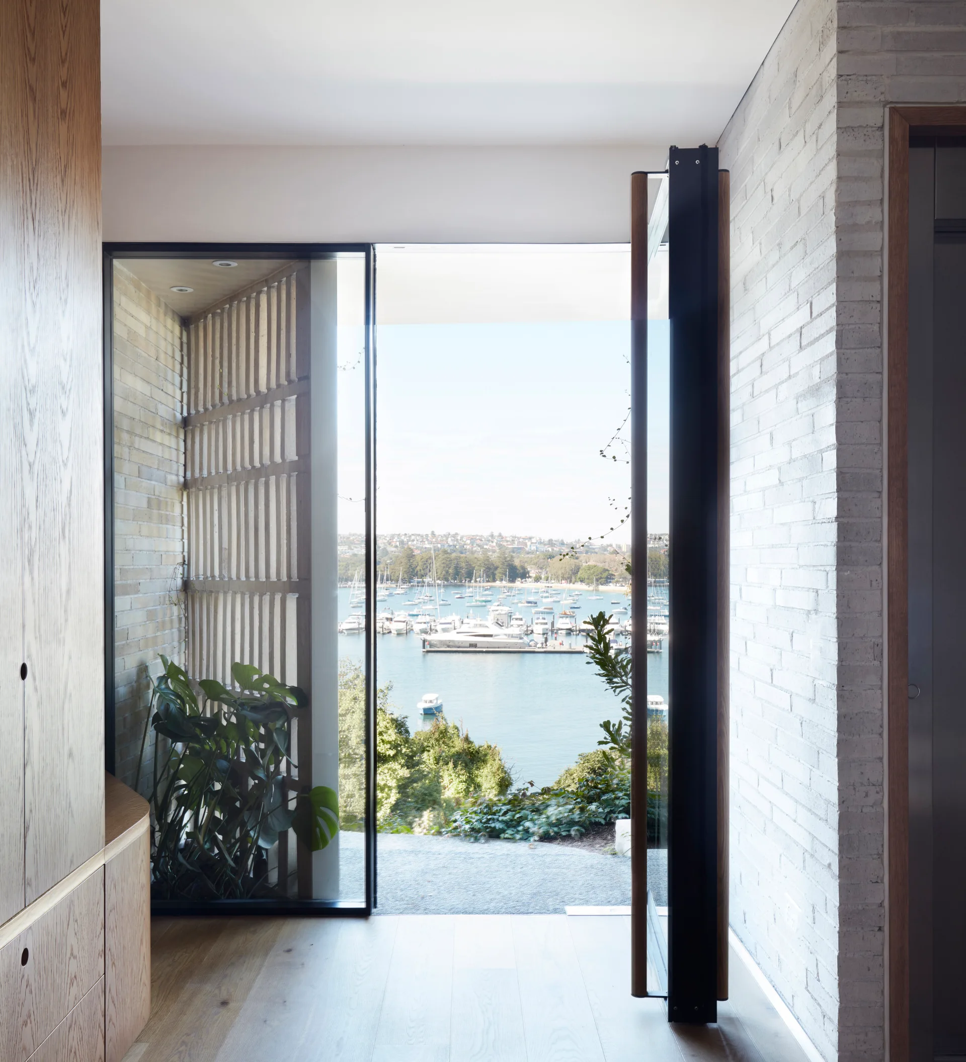 A modern entryway features a large glass pivot door opening to a harbor view, flanked by a whitewashed brick wall and oak cabinetry.