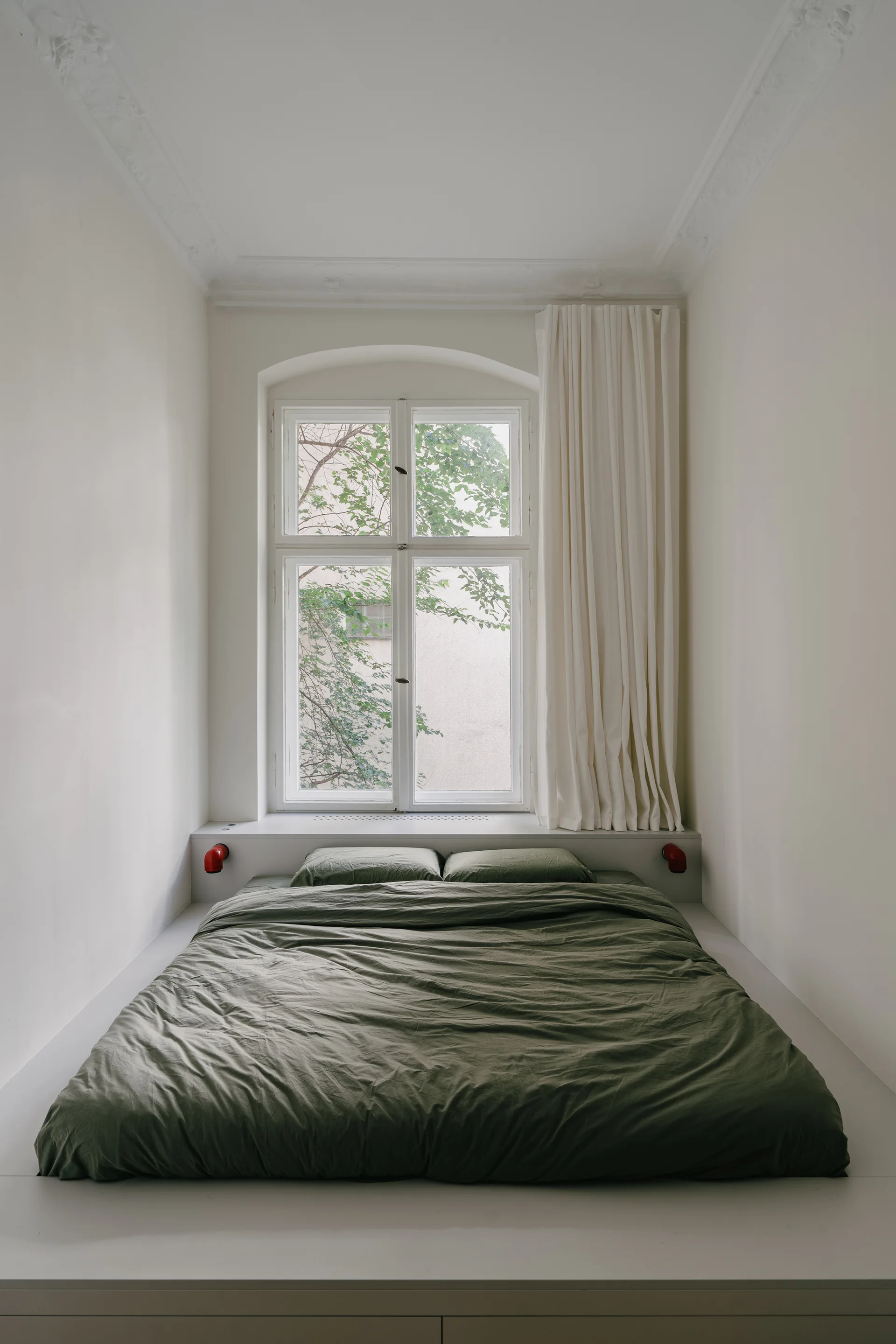 A bed with olive green bedding is positioned on a built-in white platform below a large window in a minimalist alcove bedroom.