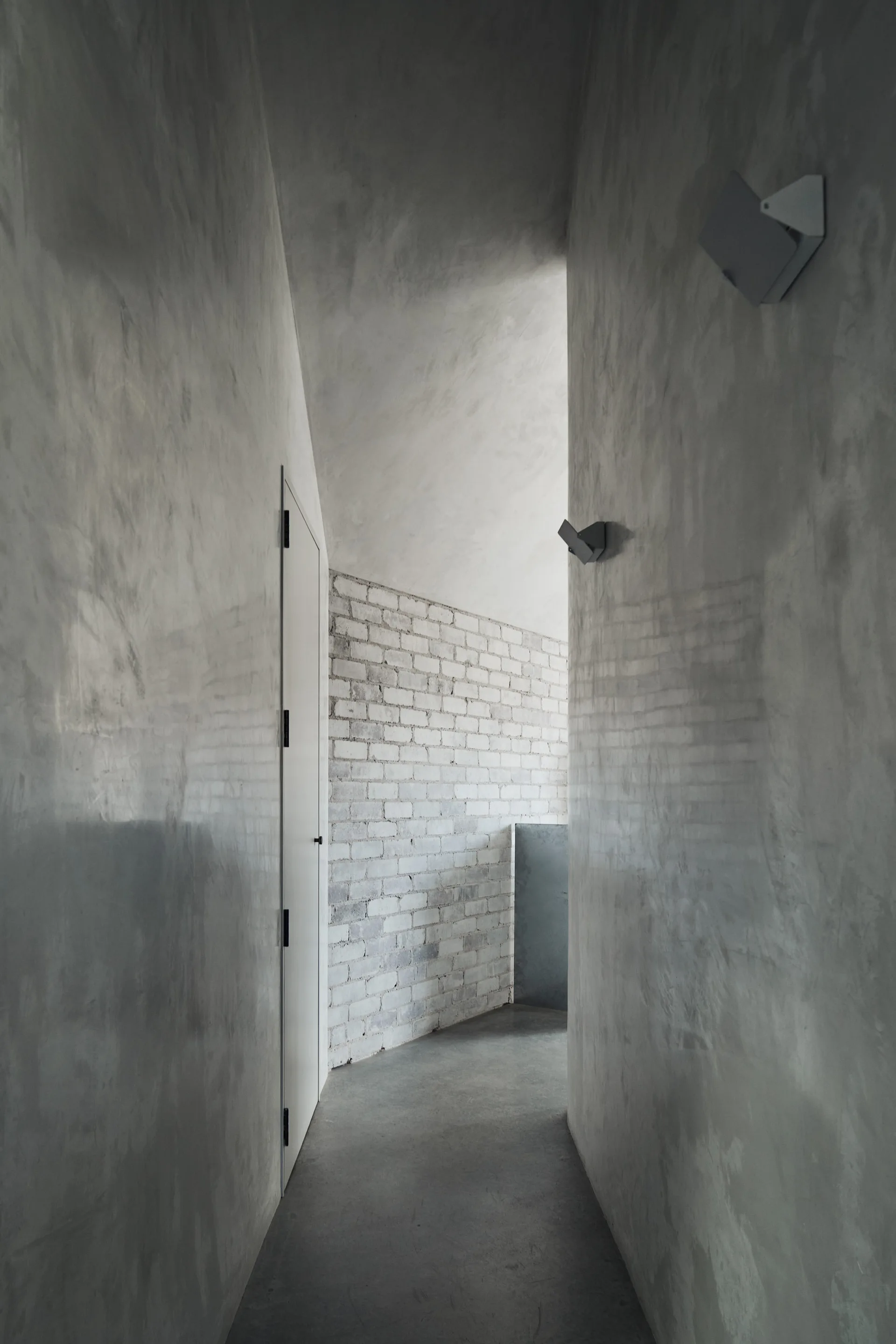A narrow minimalist hallway with smooth gray concrete walls, a flush white door, and a curved white brick wall.