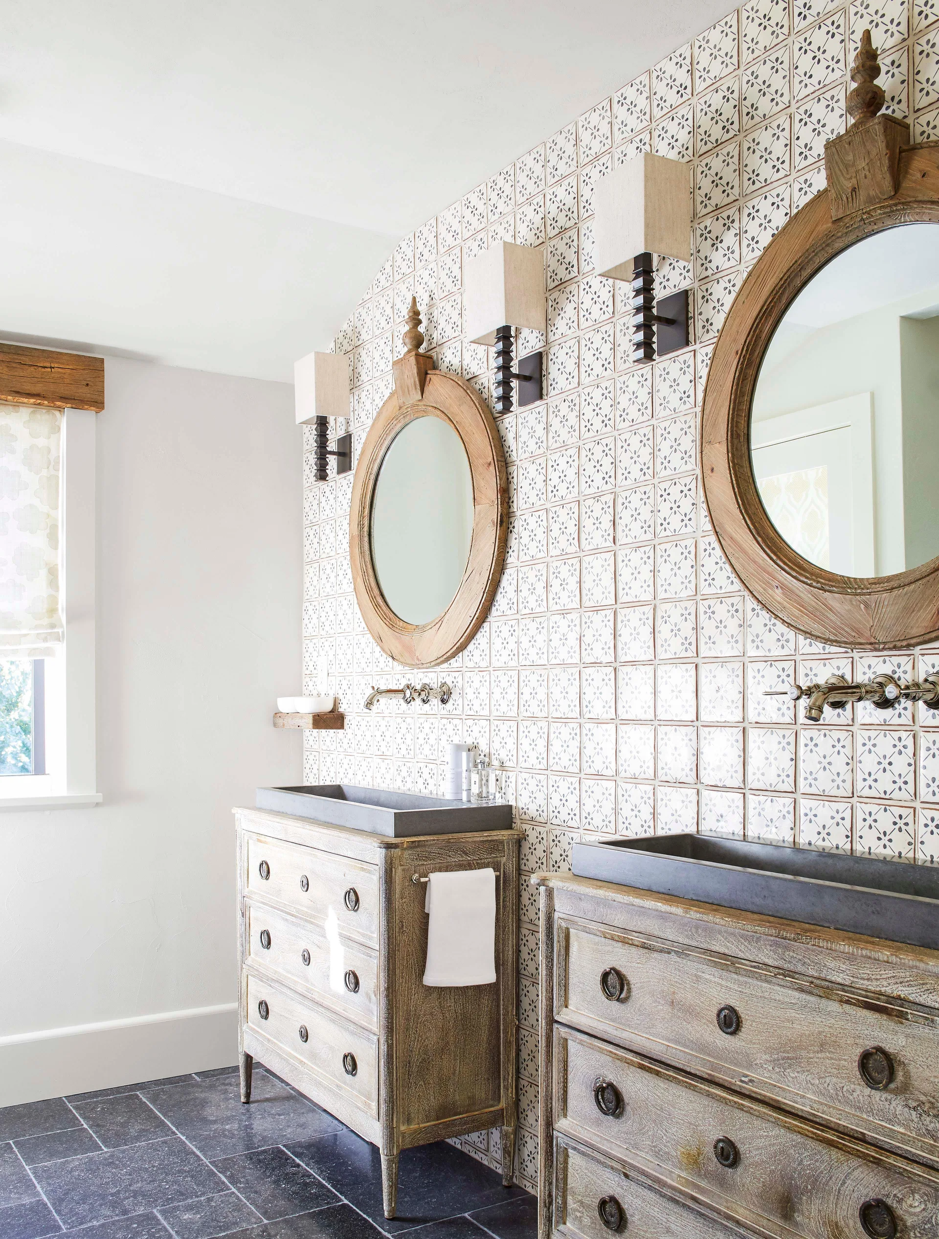 Two weathered wood dressers repurposed as bathroom vanities stand against a wall of white patterned tiles beneath round wooden mirrors.