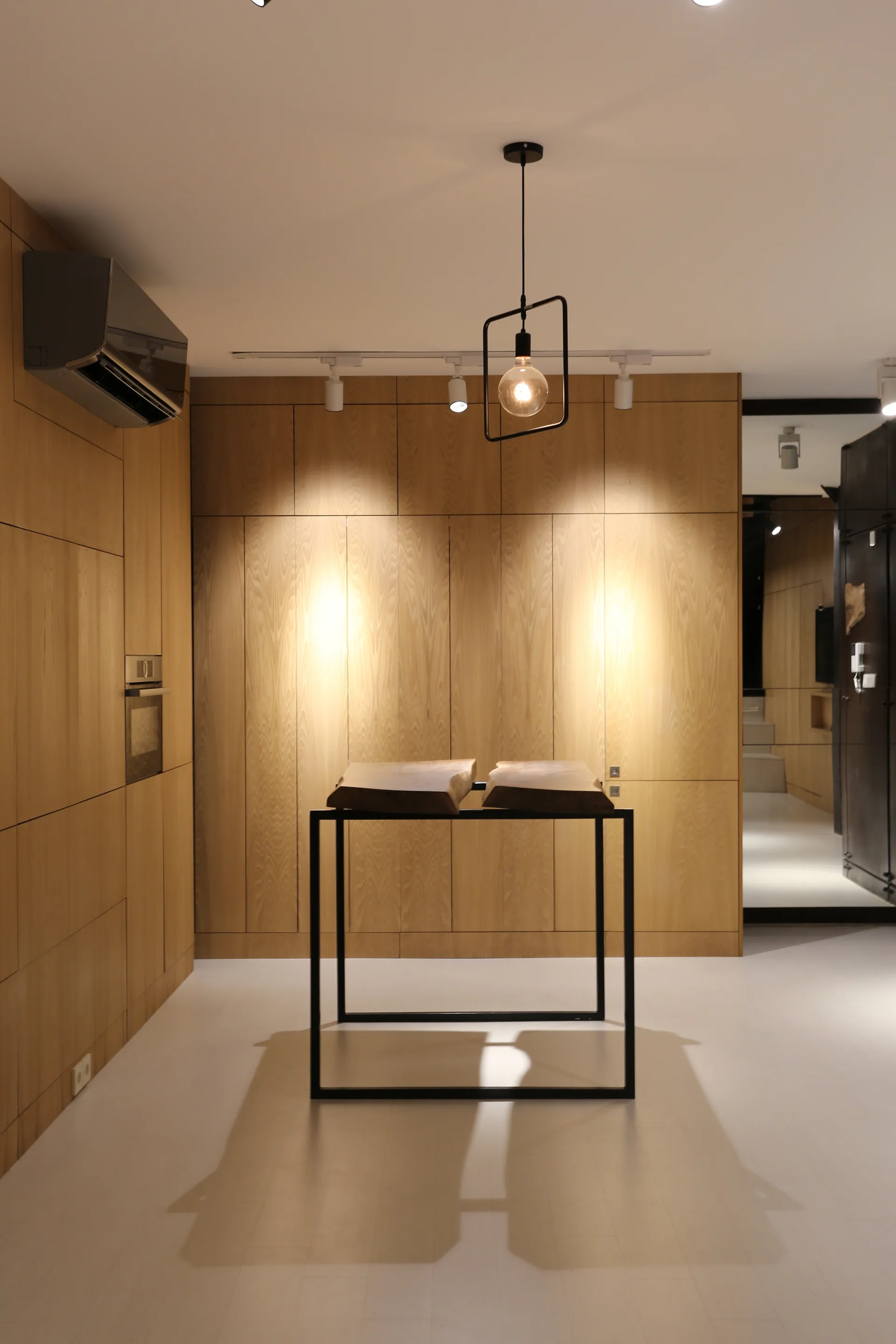 A minimalist kitchen features flush light wood cabinetry, a central table with a black metal frame, and a geometric pendant light.