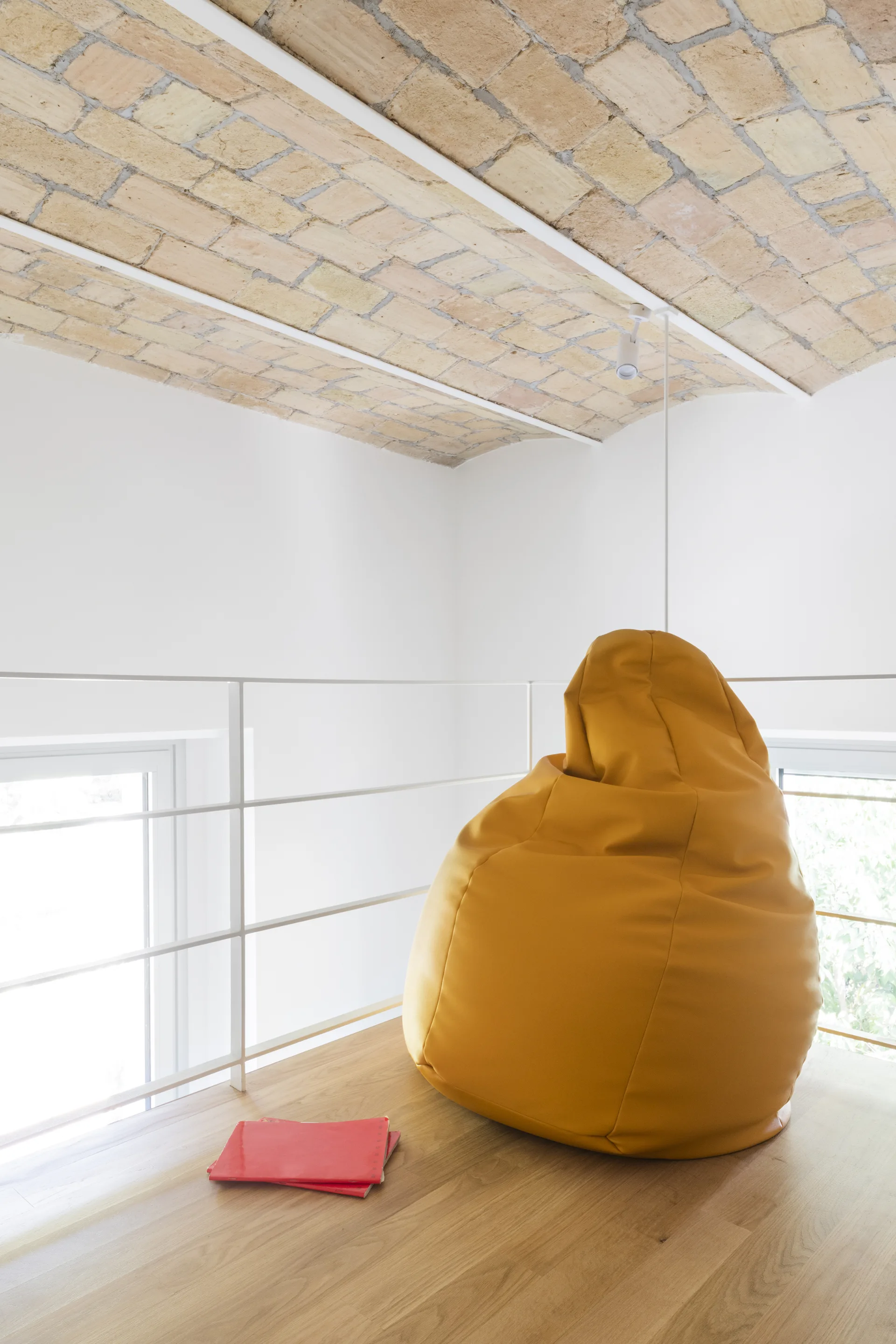 A bright lofted landing features a large yellow beanbag chair on light wood floors beneath a vaulted exposed brick ceiling with white railings.