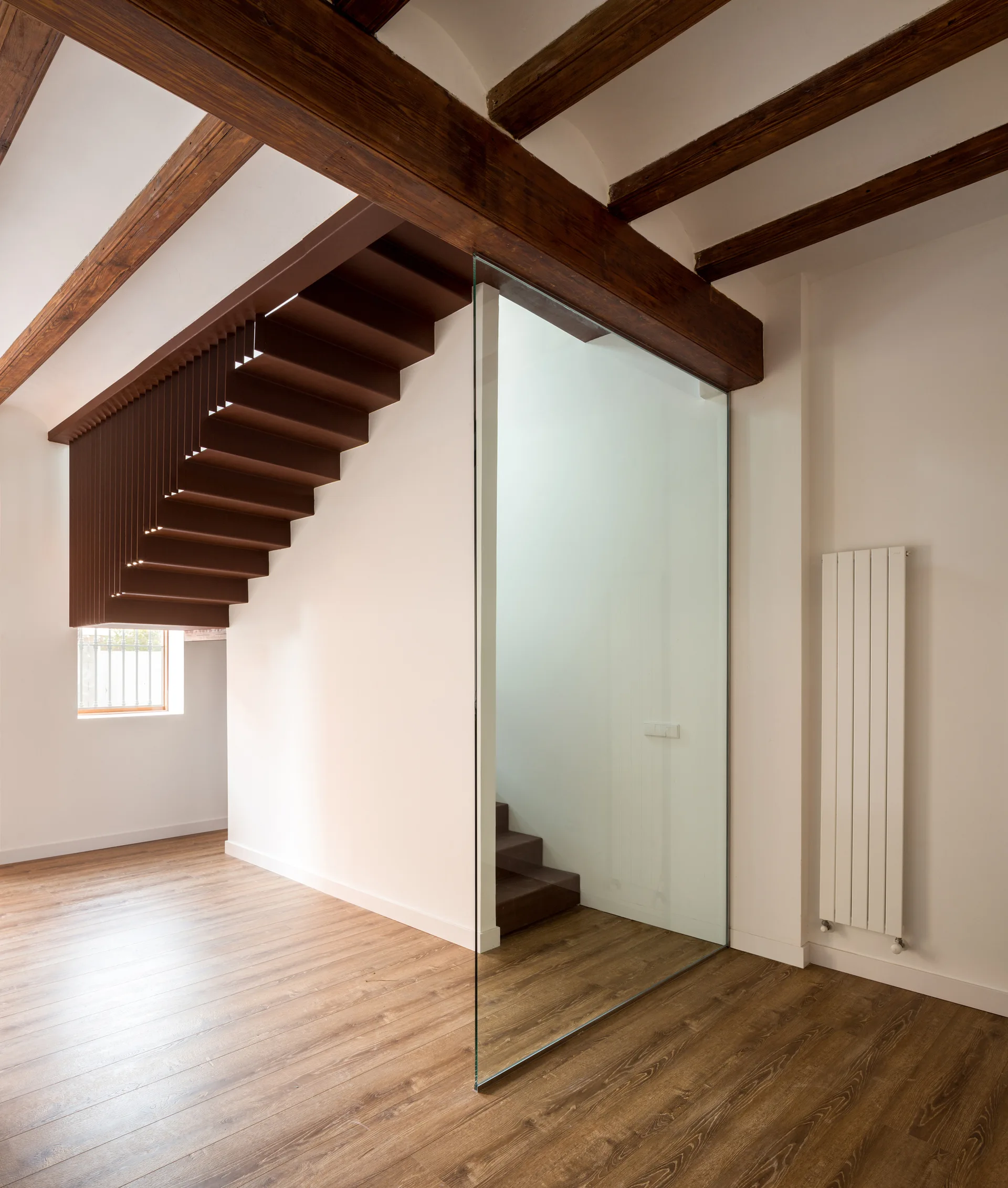 A modern interior space features a brown metal suspended staircase, a frameless glass partition, exposed wood ceiling beams, and wood floors.