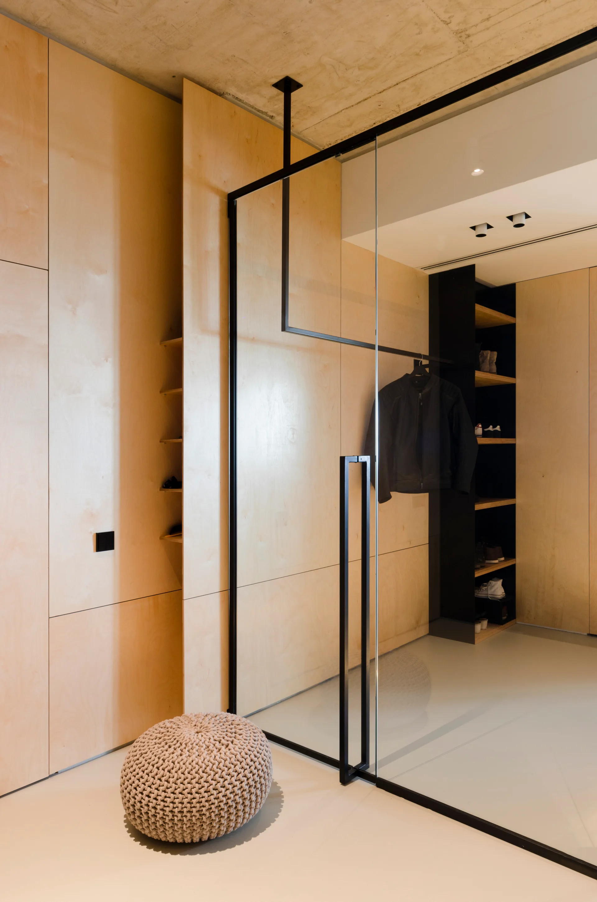 A minimalist dressing room featuring built-in light wood cabinetry, a black-framed glass partition, a knitted beige pouf, and a raw concrete ceiling.