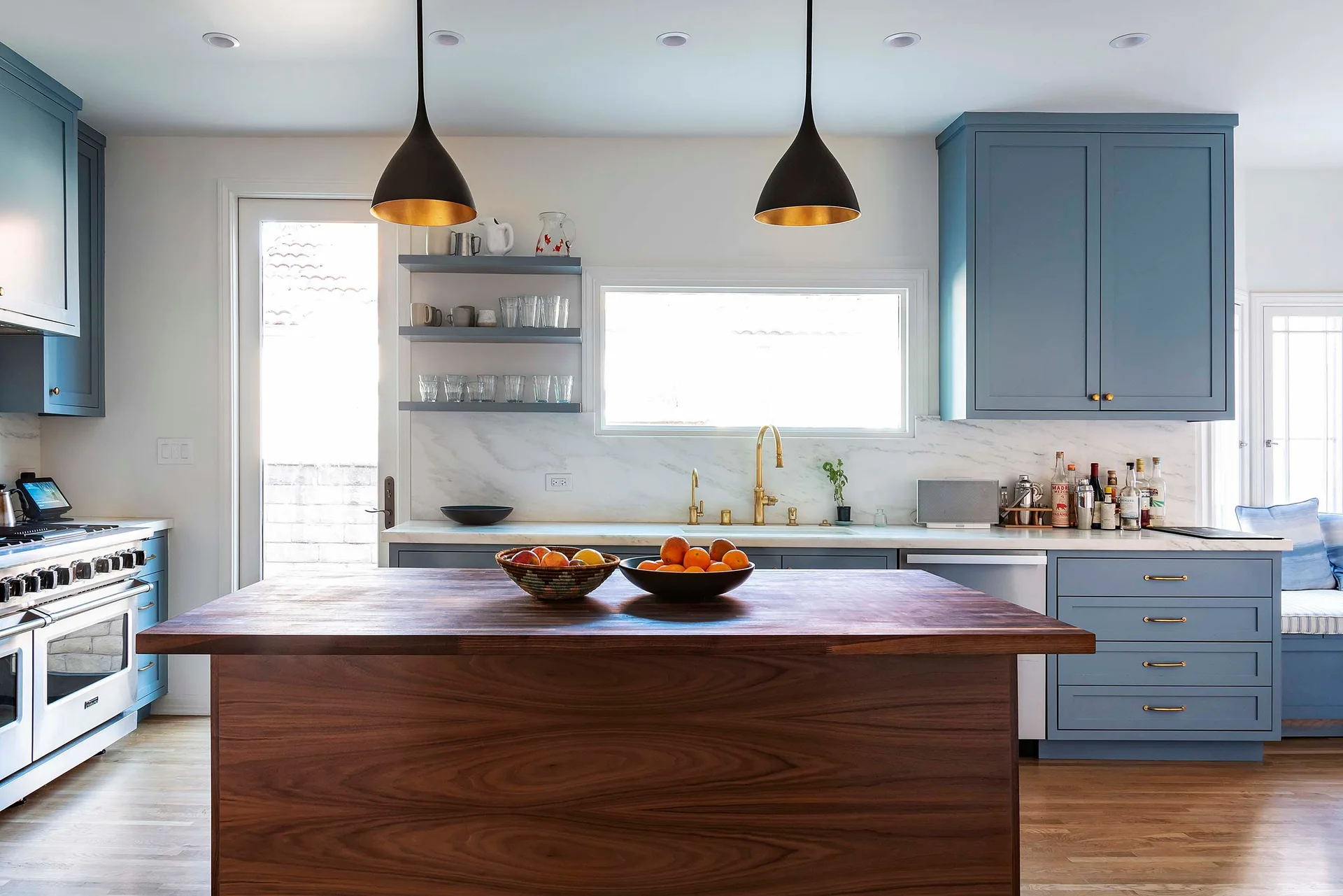 A bright kitchen features muted blue cabinets, a rich walnut wood island, white marble counters, and black dome pendant lights.