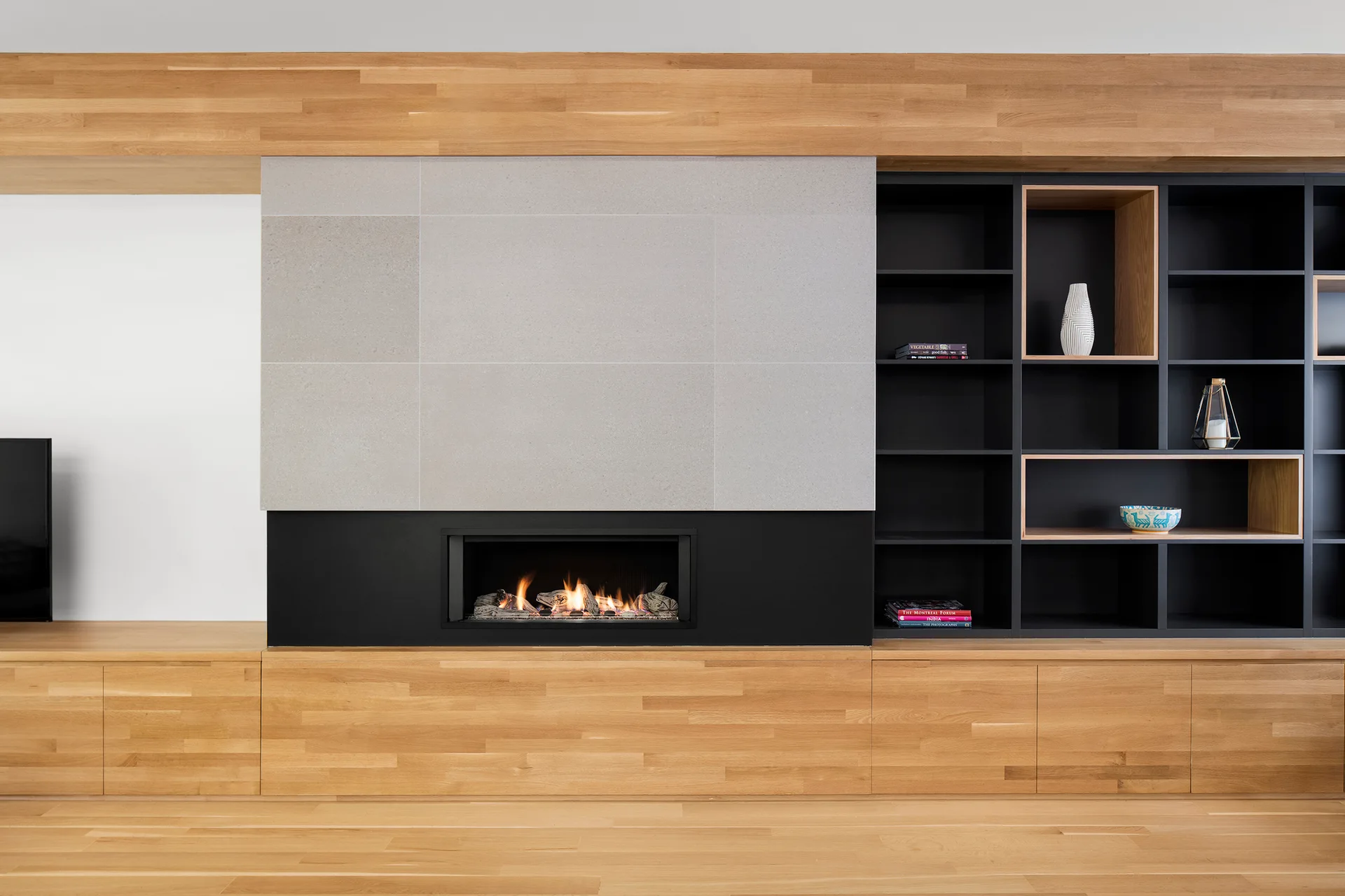A modern linear fireplace set in a matte black surround, topped with grey stone panels and flanked by light wood cabinetry and black open shelving.