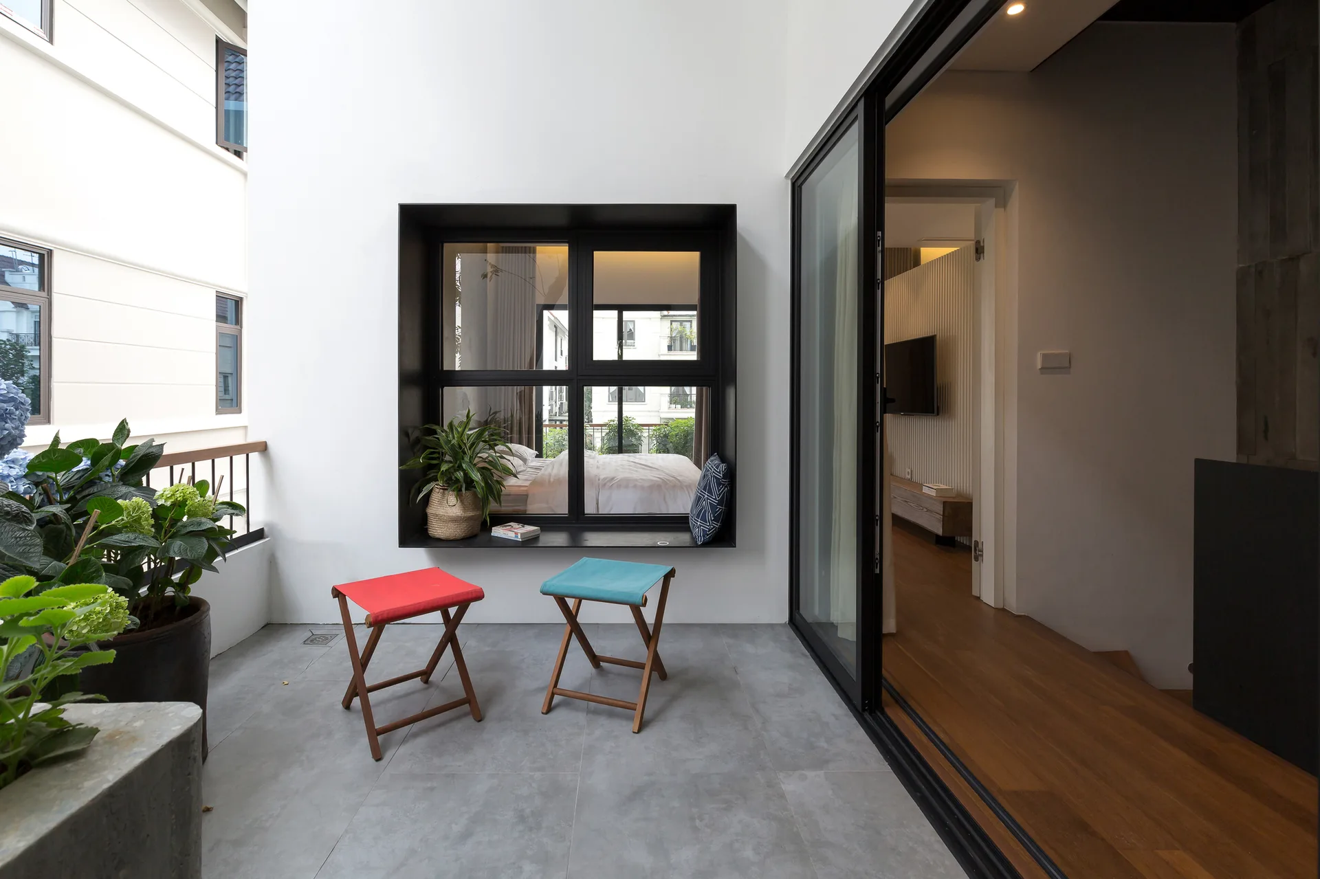 A modern outdoor balcony features gray tile flooring, two folding stools, and a large black-framed window looking into a bedroom.