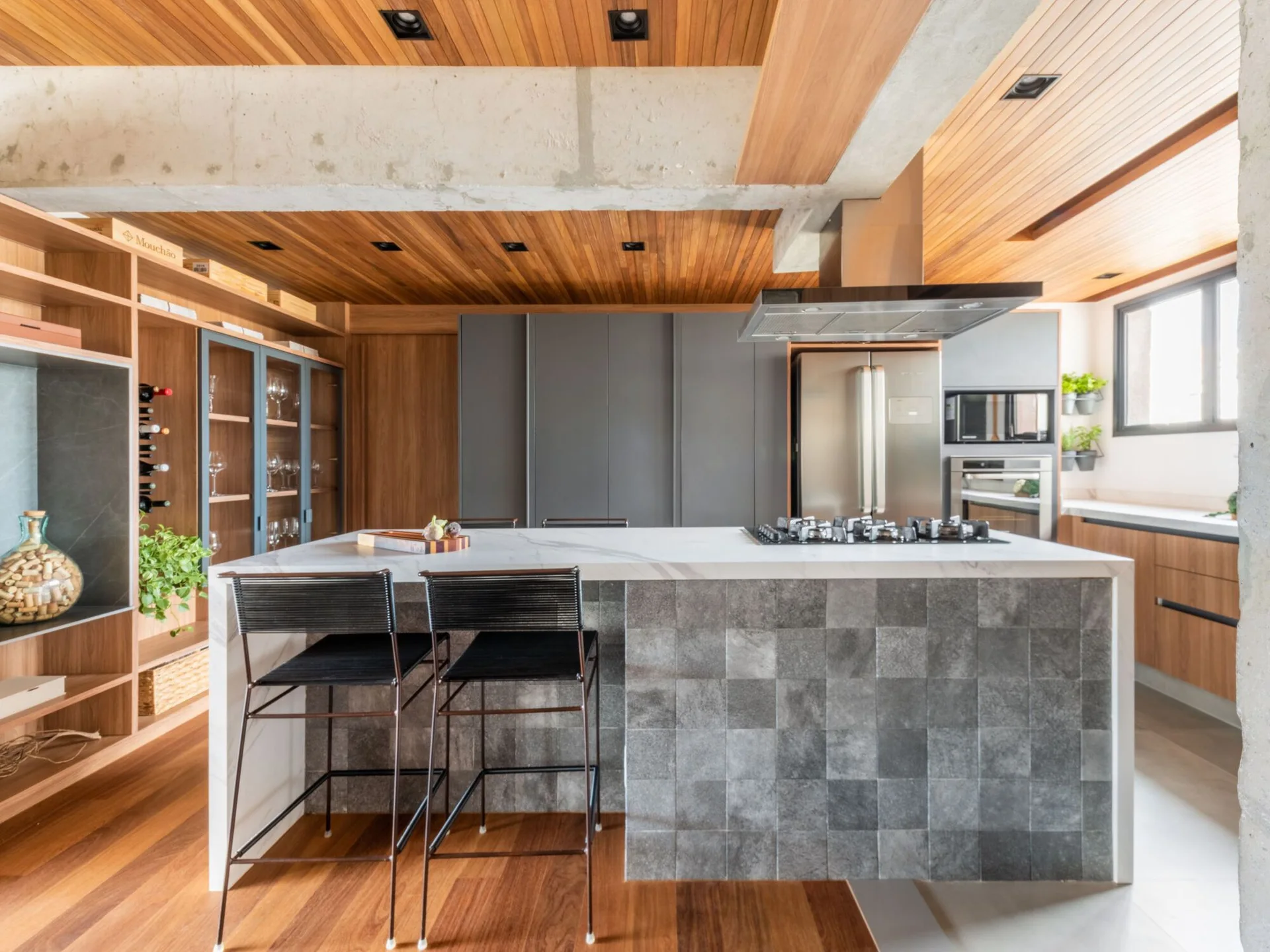A contemporary kitchen features a wood slat ceiling, an exposed concrete beam, and a marble-topped island with a textured grey tiled front.