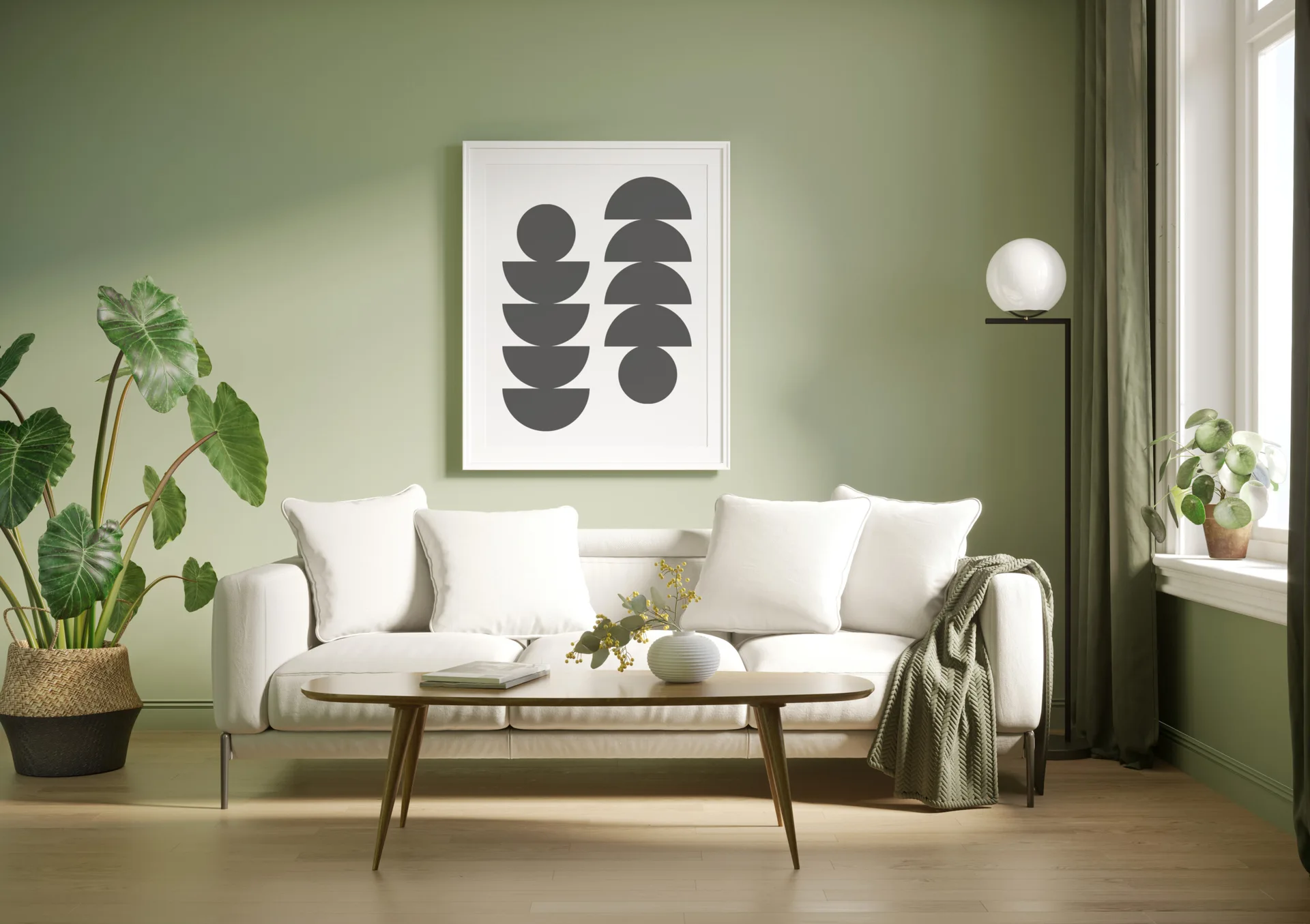 A minimalist living room features a white sofa on wood floors against a sage green wall with framed abstract art and a large leafy potted plant.