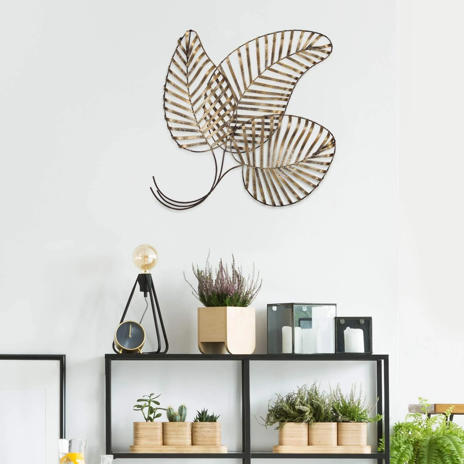 A gold leaf metal wall sculpture hangs on a white wall above a black display shelf styled with potted plants and a small table lamp.