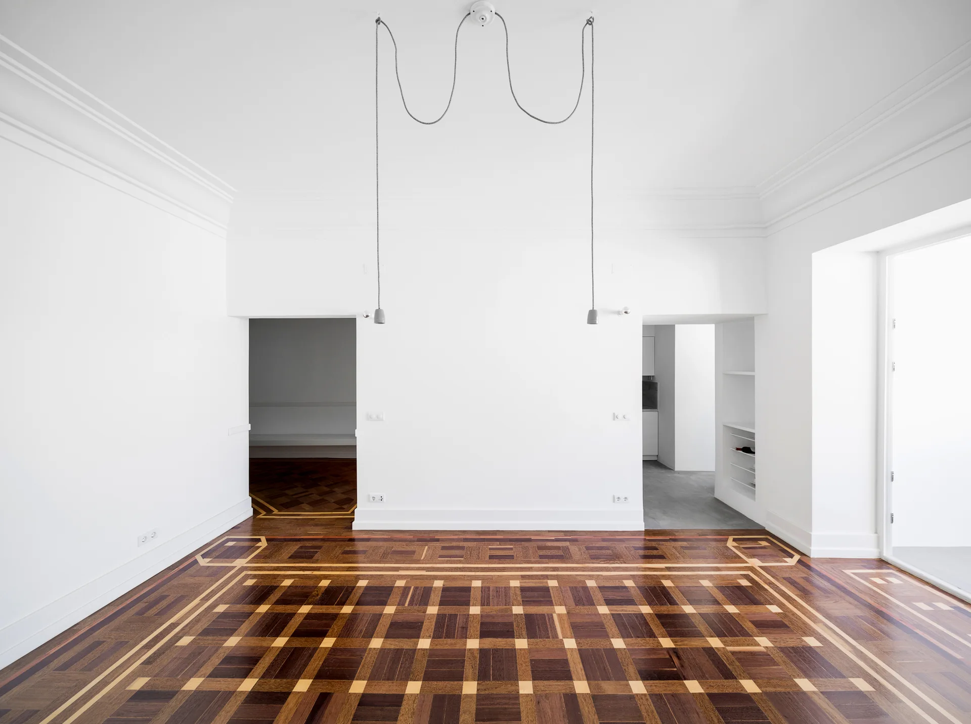 An empty room with stark white walls, ornate geometric parquet wood flooring, crown molding, and two draped black pendant light cords hanging from the ceiling.