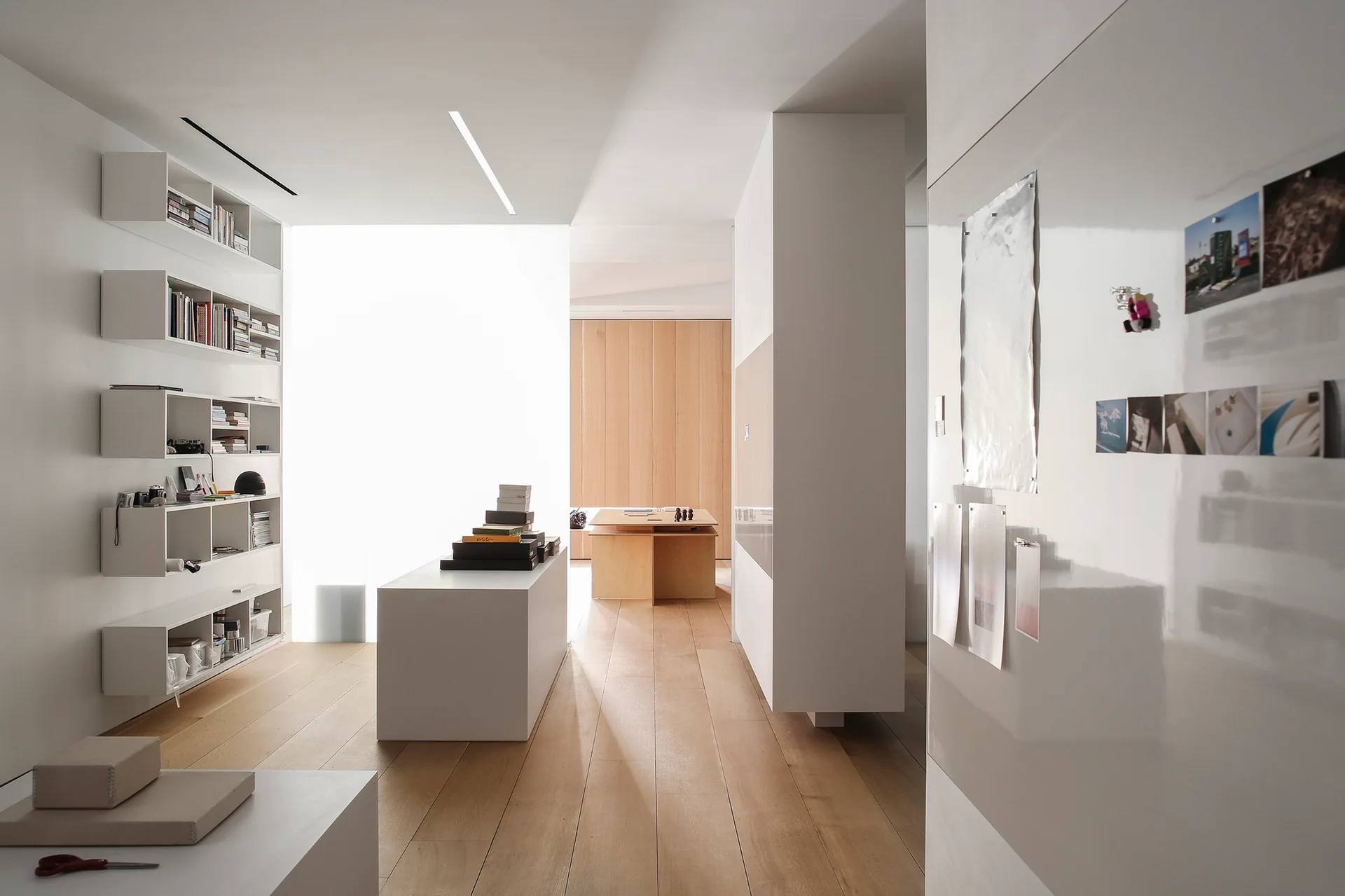 A minimalist home studio features wall-mounted white box shelving, a central white island desk, and light wood flooring.