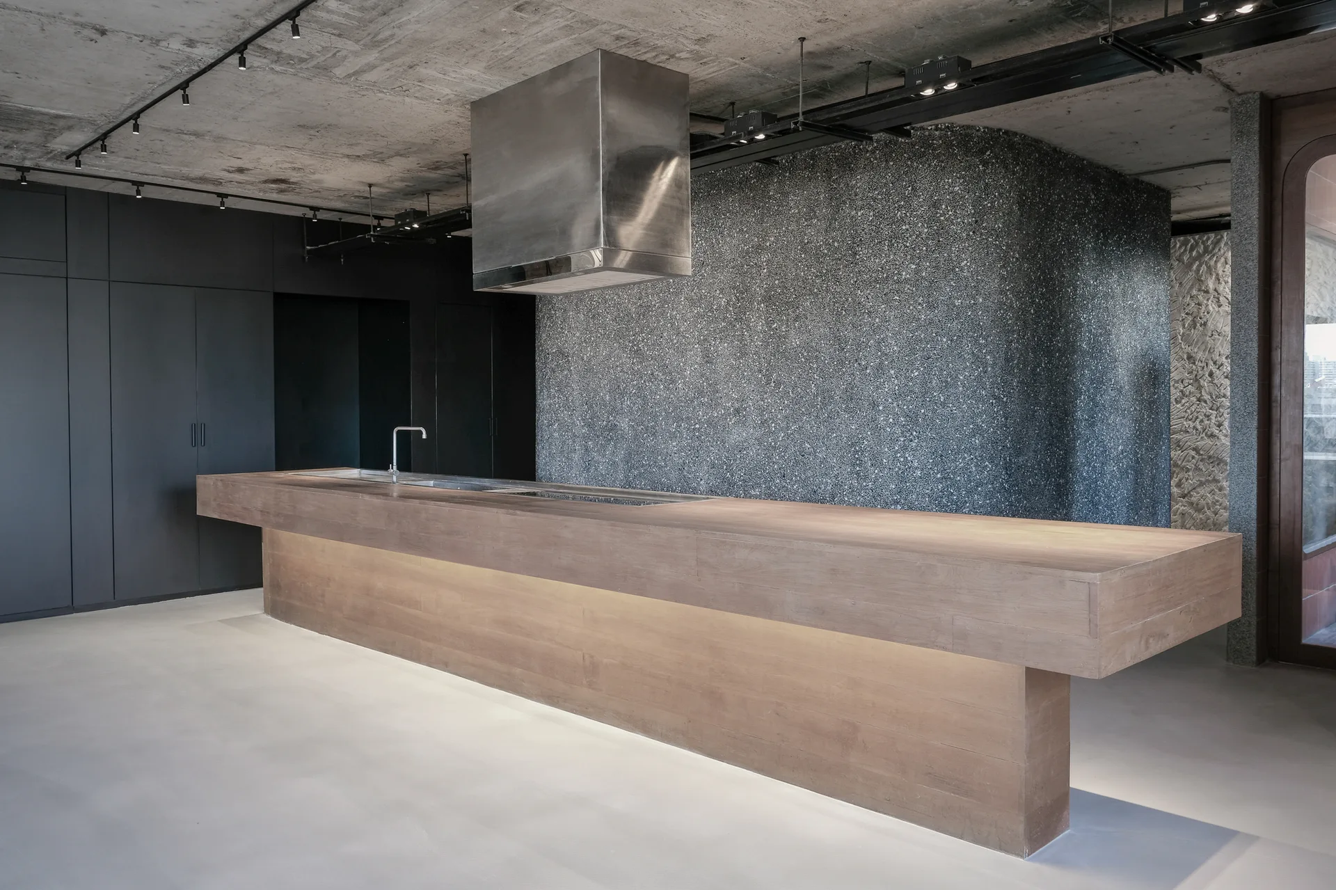A minimalist kitchen features a long wooden island, a stainless steel range hood, black cabinets, and a curved gray textured terrazzo wall.