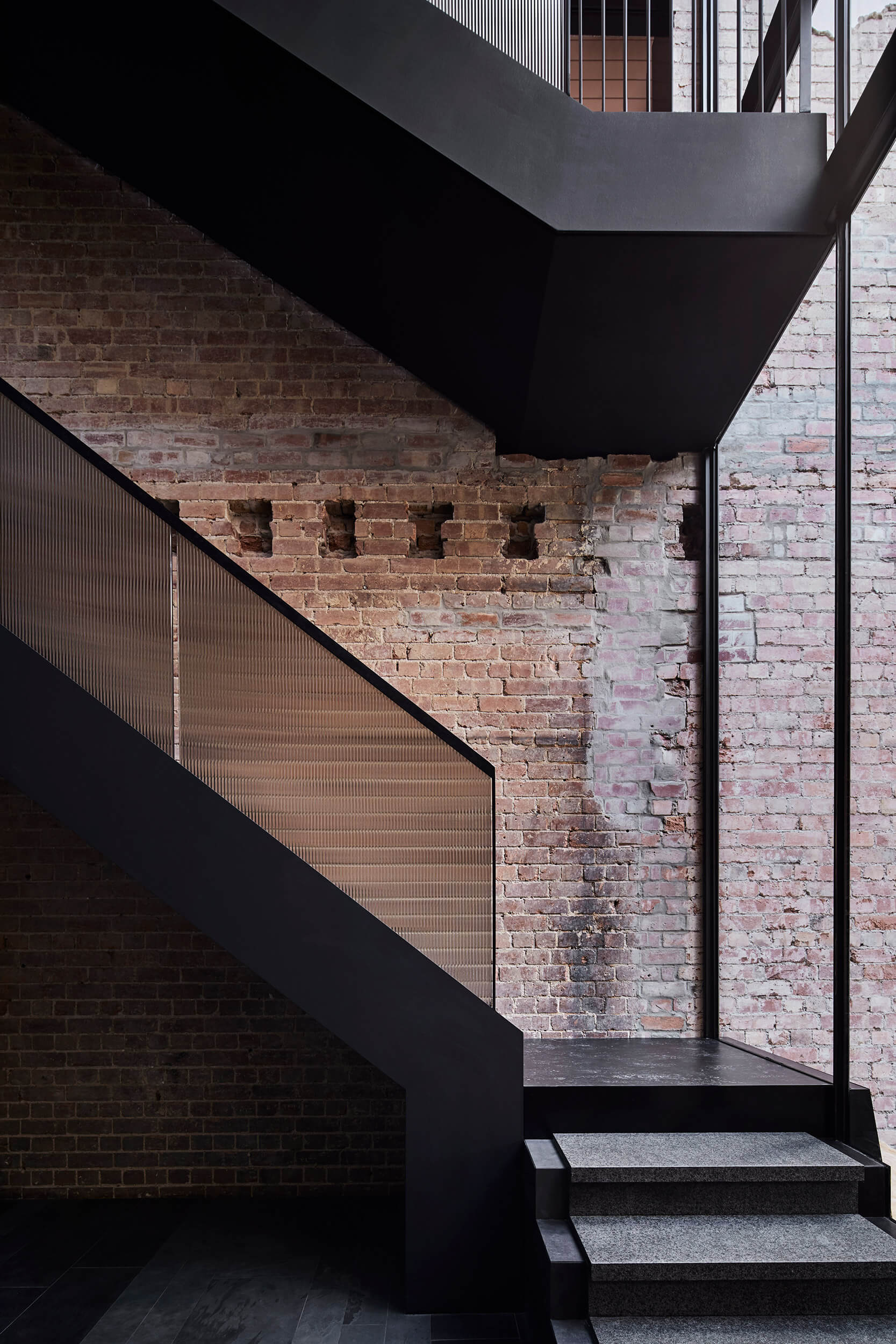 A matte black metal staircase features a ribbed glass railing against an old exposed red brick wall.