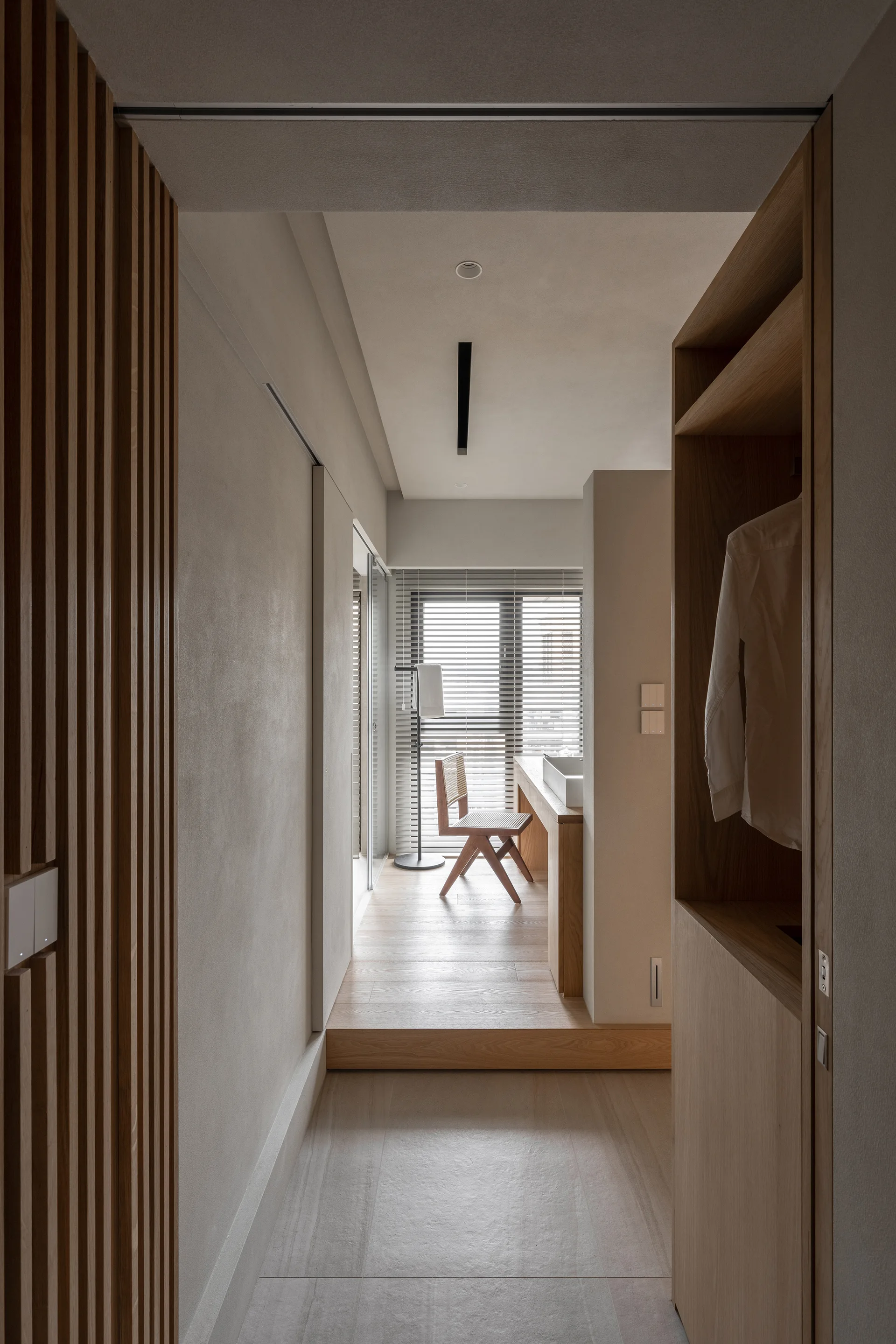 A minimalist dressing area featuring built-in oak wardrobes, a raised wooden platform floor, a vanity table with a cane chair, and venetian blinds.