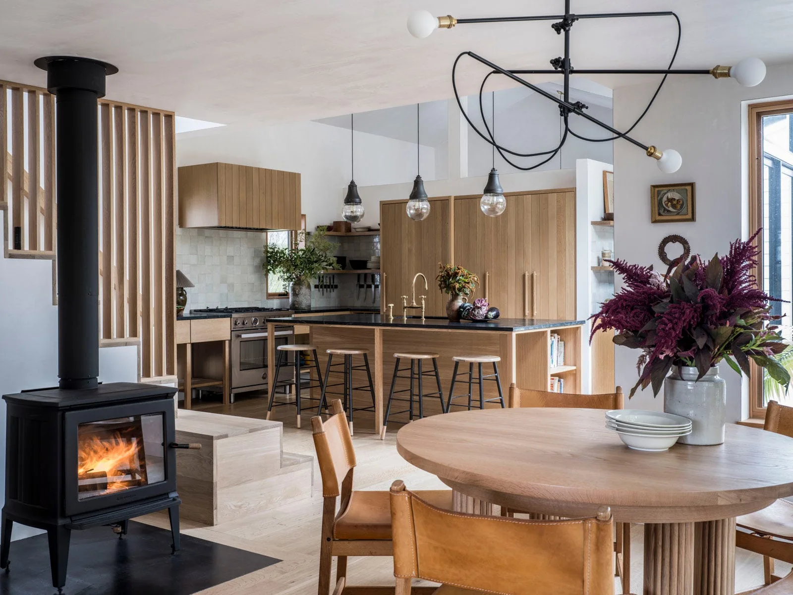 An open-concept kitchen features natural oak cabinetry, a round wooden dining table with leather chairs, and a black wood-burning stove.