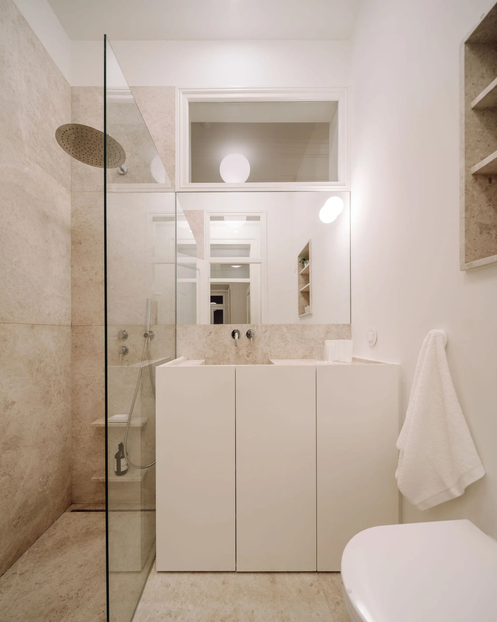 A modern bathroom featuring beige stone tile walls, a glass walk-in shower, a white vanity cabinet, and a rectangular transom window above the mirror.