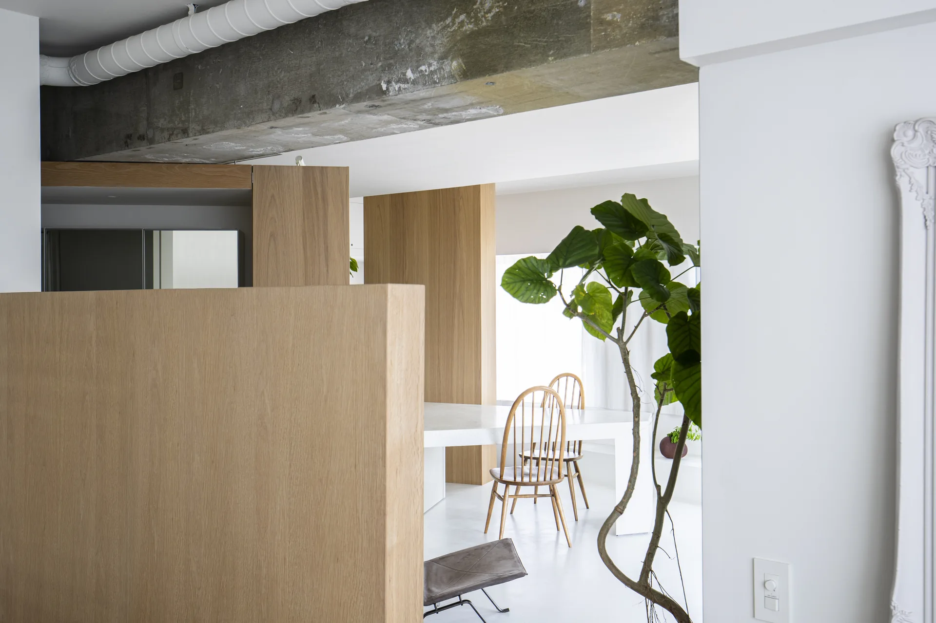 A minimalist dining room with a white table, wooden spindle-back chairs, light wood partition walls, and an exposed concrete ceiling beam.