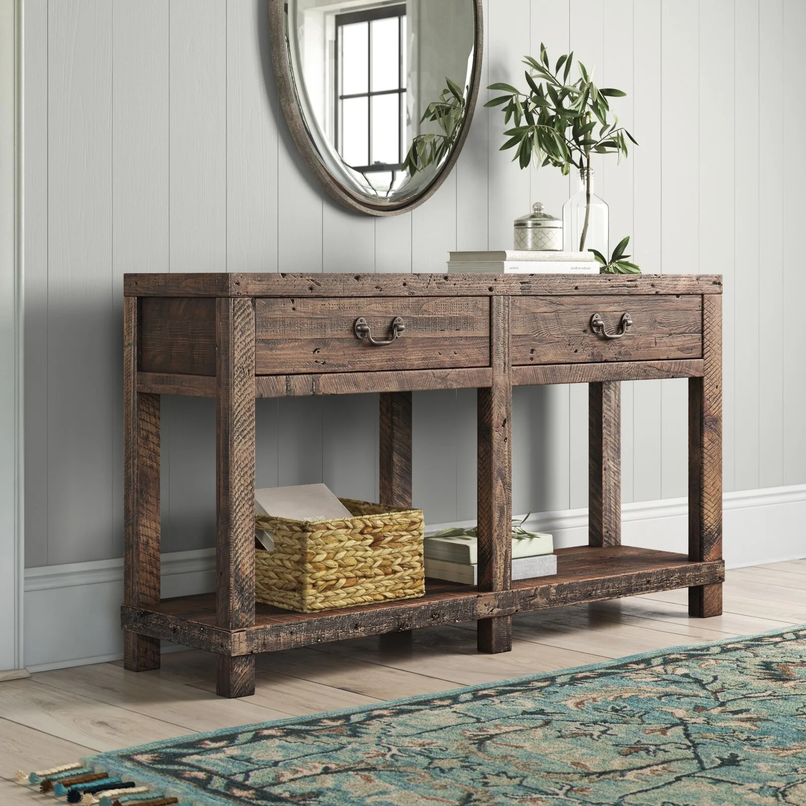 A rustic wooden console table with two drawers stands against a light grey vertical paneled wall under an oval mirror.