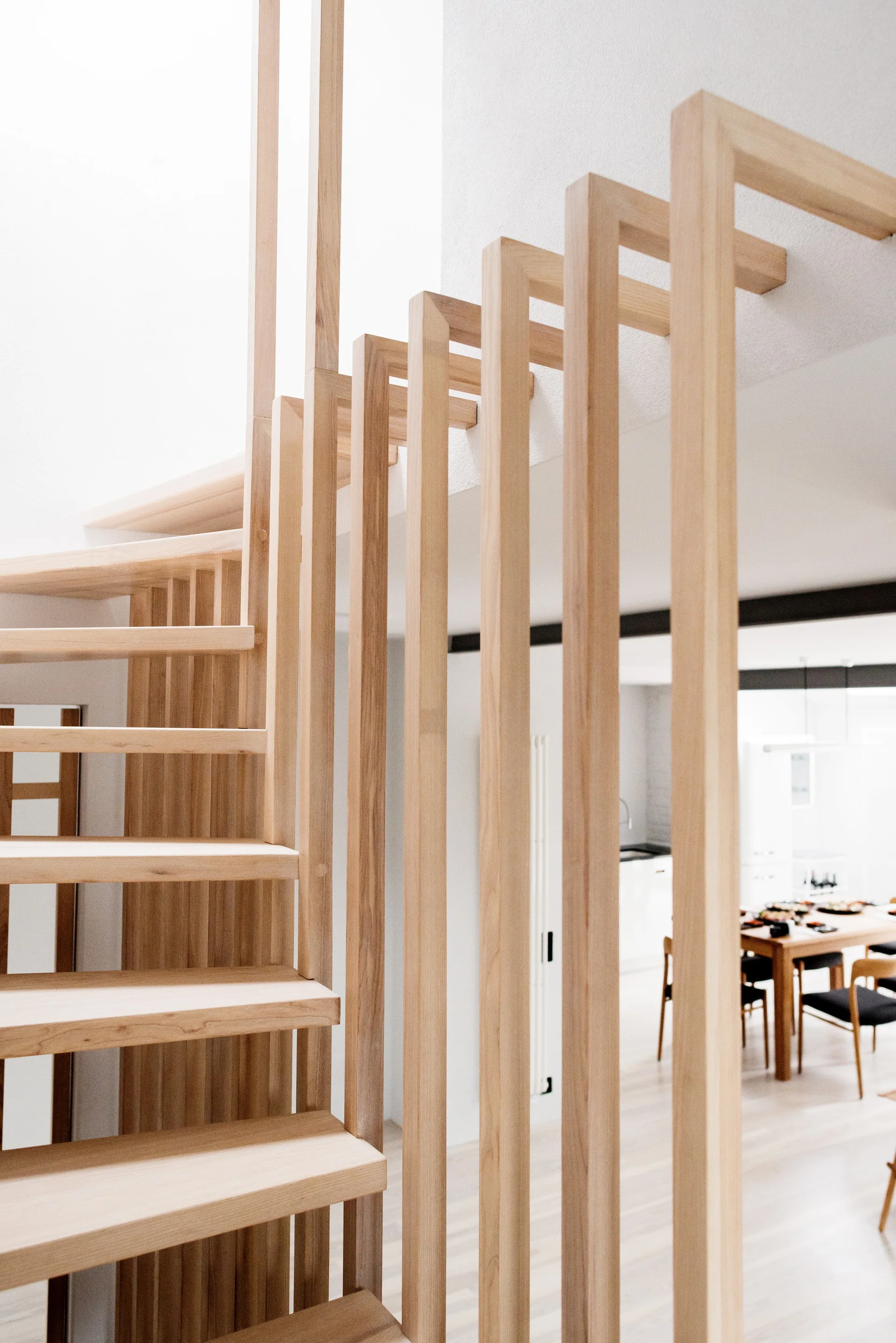 A light wood staircase with a tall vertical slat railing overlooks an open-plan dining room with a wooden table and chairs.