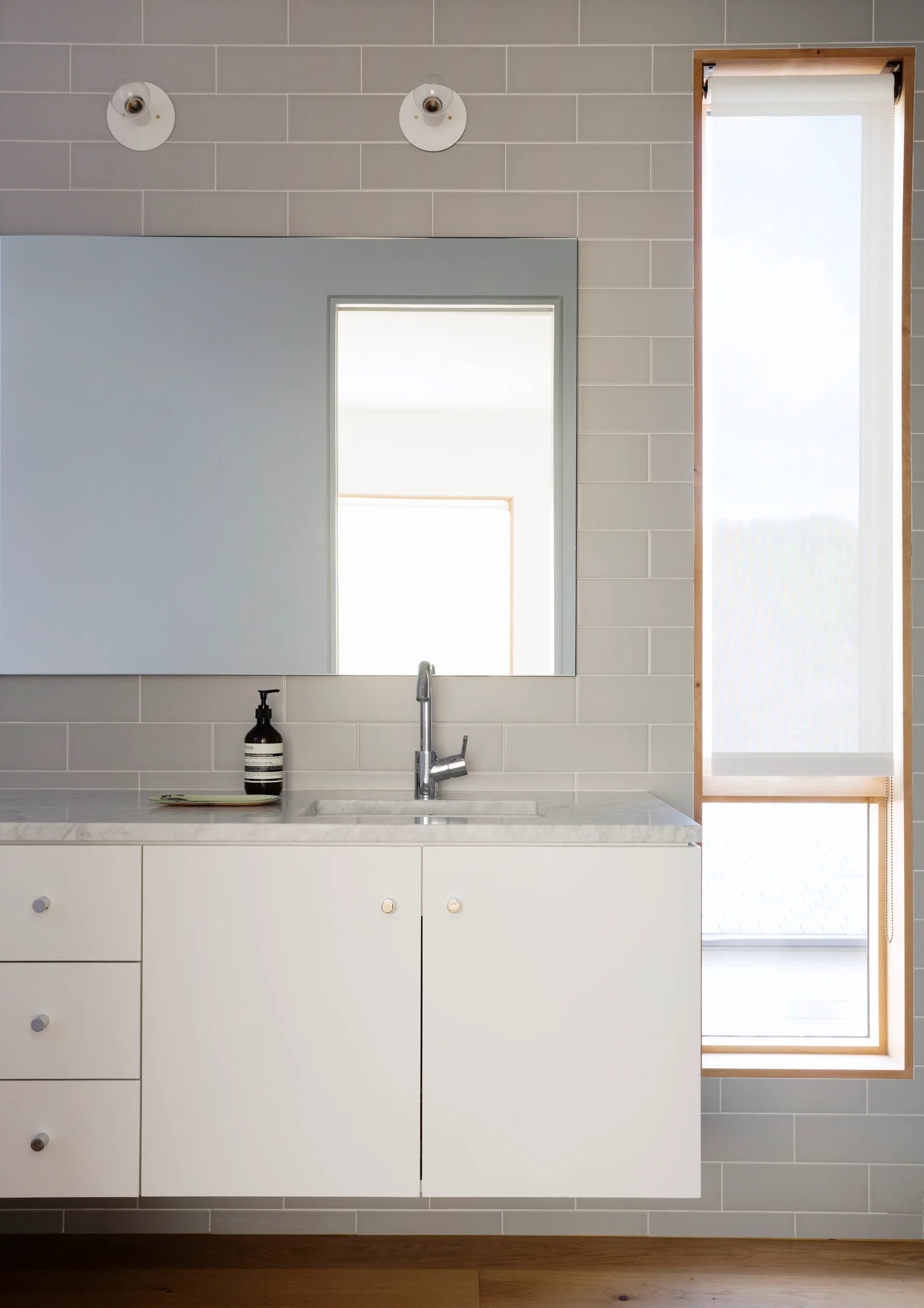 A modern bathroom vanity features a floating white cabinet with a marble top, gray subway tiled walls, and a tall vertical window with wood trim.