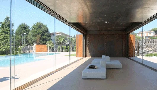 Low white modular seating sits inside a glass-walled lounge featuring a dark metal ceiling and views of an outdoor pool.