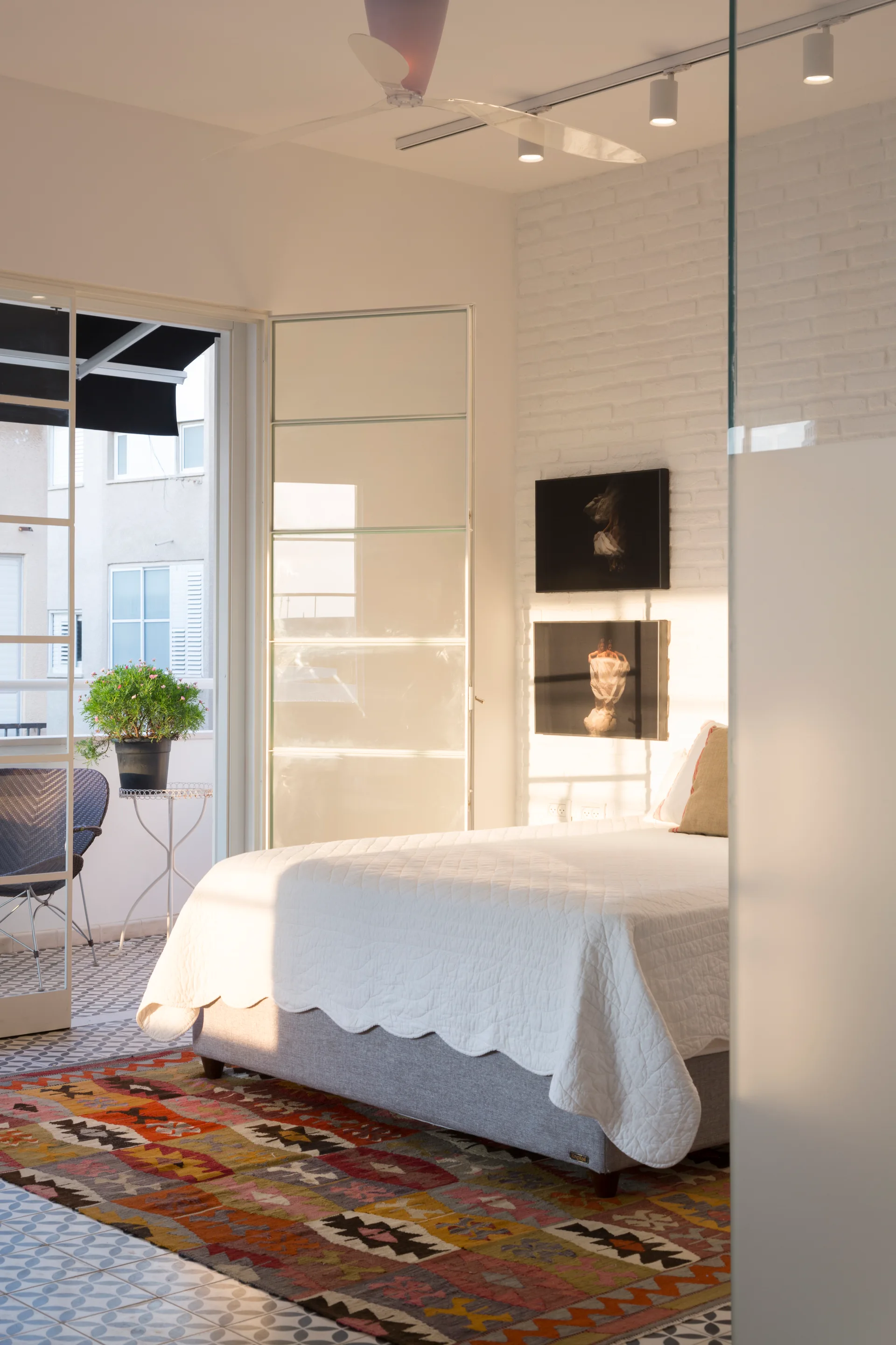 A sunlit bedroom with a quilted bed next to a white brick wall and a colorful patterned rug, opening onto a tiled balcony.