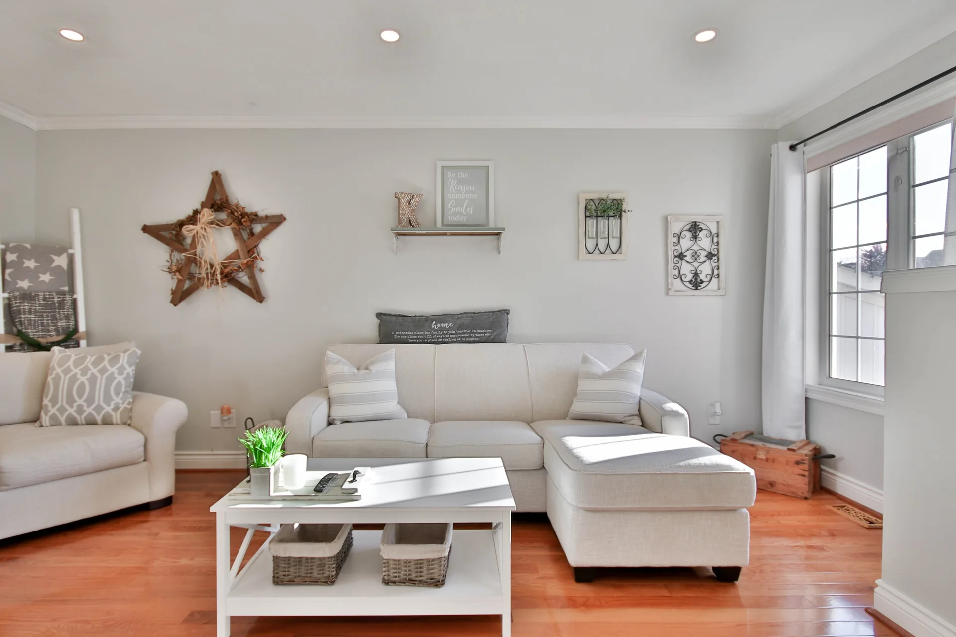 A bright living room features a cream sectional sofa, a white coffee table with baskets, and rustic wooden star wall decor on a light grey wall.