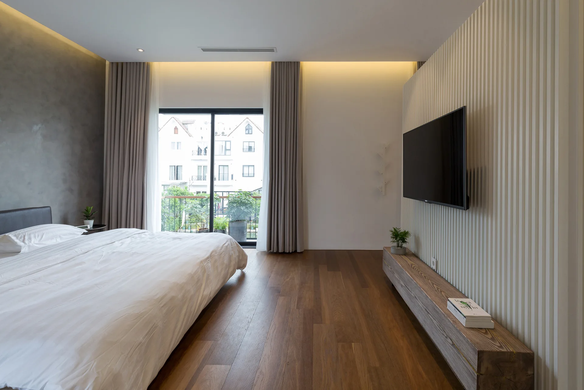 A minimalist bedroom features a bed with white linens, a wall-mounted TV on a white fluted accent wall, and a low wooden block media console.