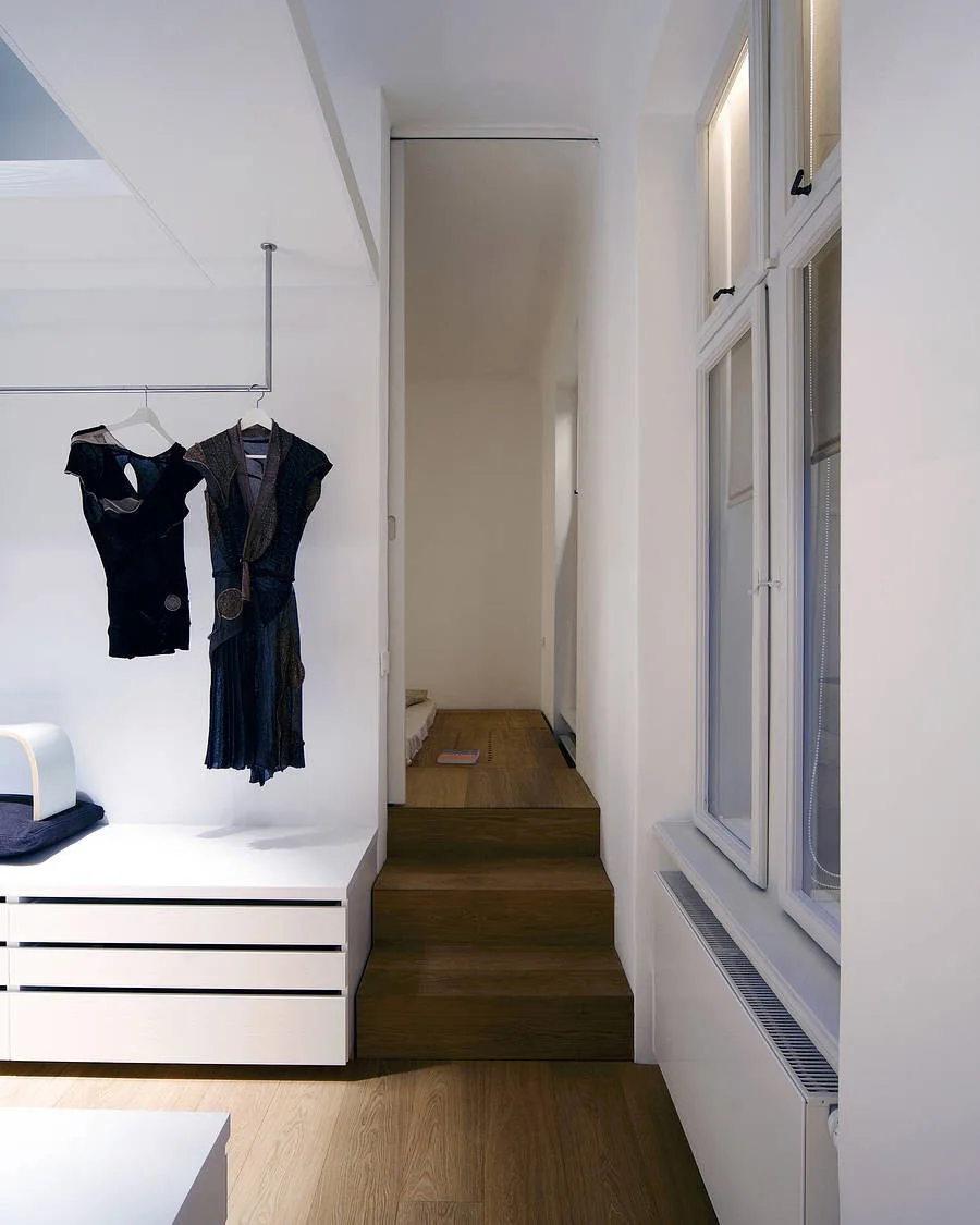 A minimalist white dressing area featuring low chest drawers, a ceiling-mounted clothing rail, and short wooden steps leading to an upper floor.