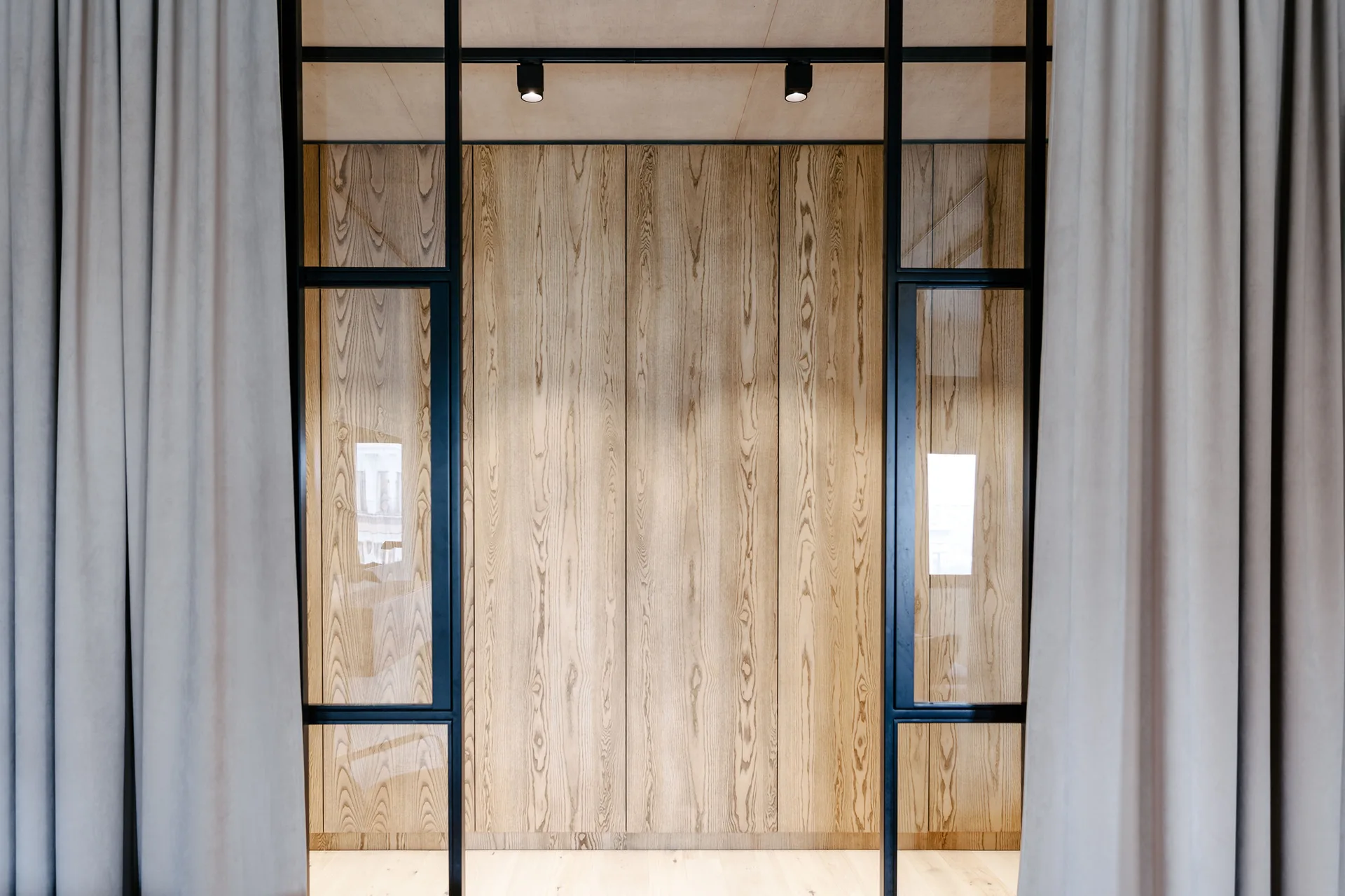 An interior view of a light wood paneled wall framed by black metal glass partition doors and soft grey drapes.
