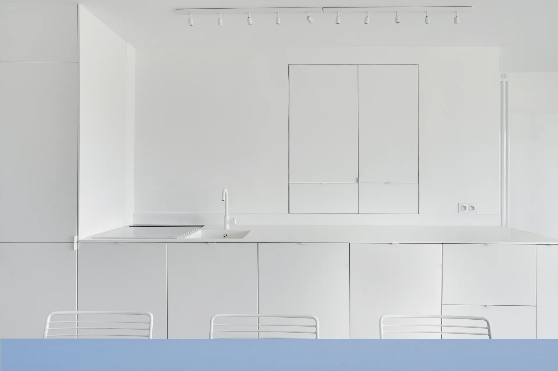 A minimalist white kitchen features smooth handleless cabinets, an integrated sink with a white faucet, and matching white wire chairs behind a blue tabletop.