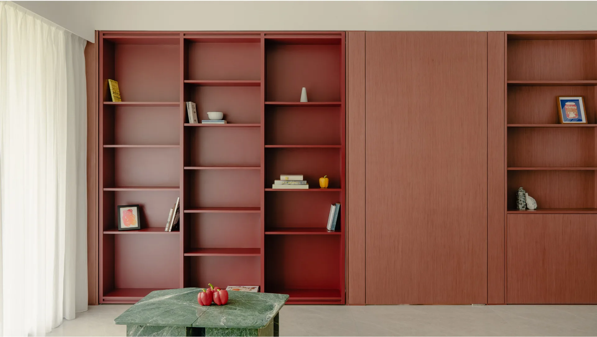 A minimalist room features a large terracotta red built-in shelving unit next to a matching wood-paneled wall and a green marble coffee table.