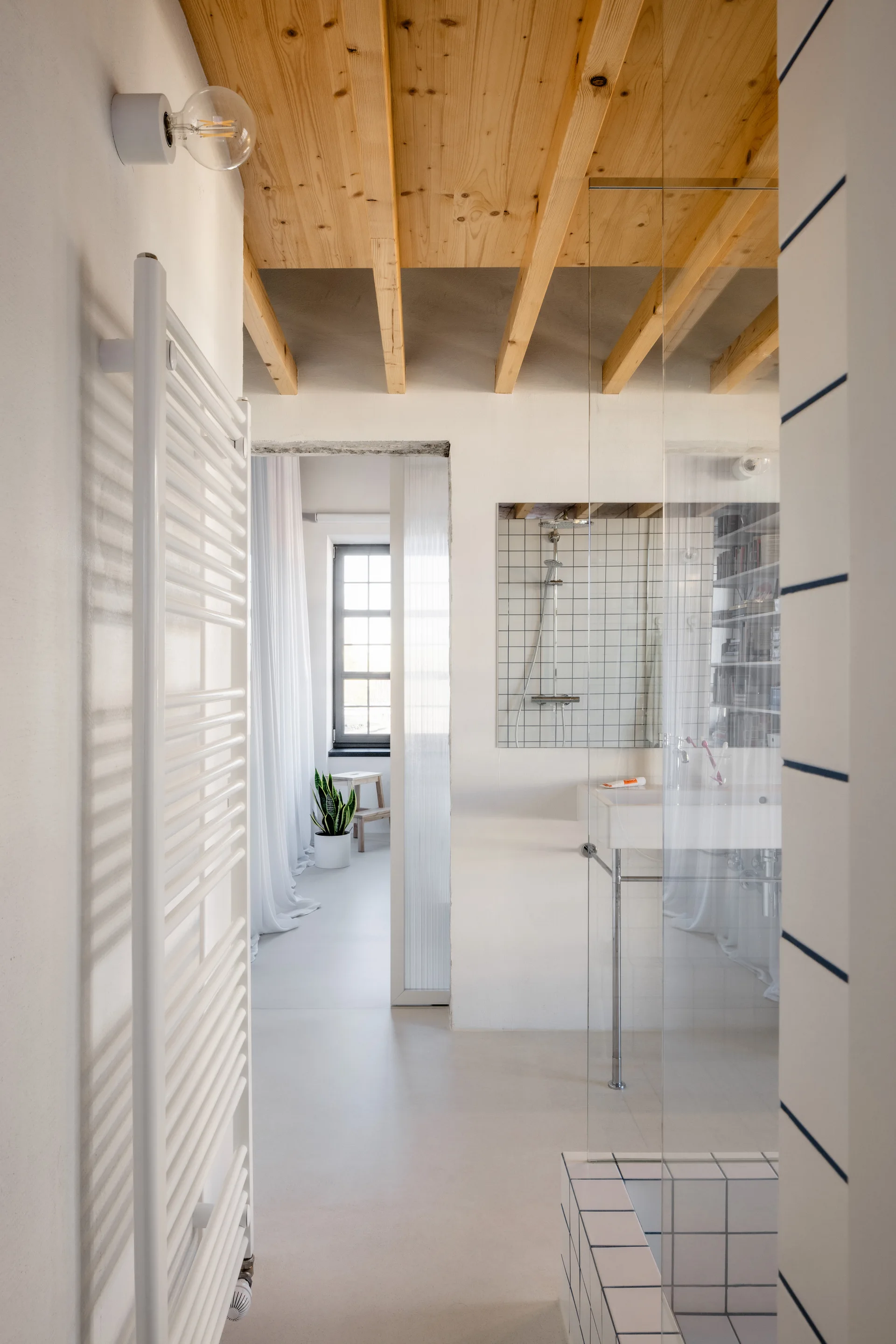 A minimalist white bathroom featuring exposed light wood ceiling beams, a wall-mounted sink, a grid-tiled shower, and a white heated towel rack.