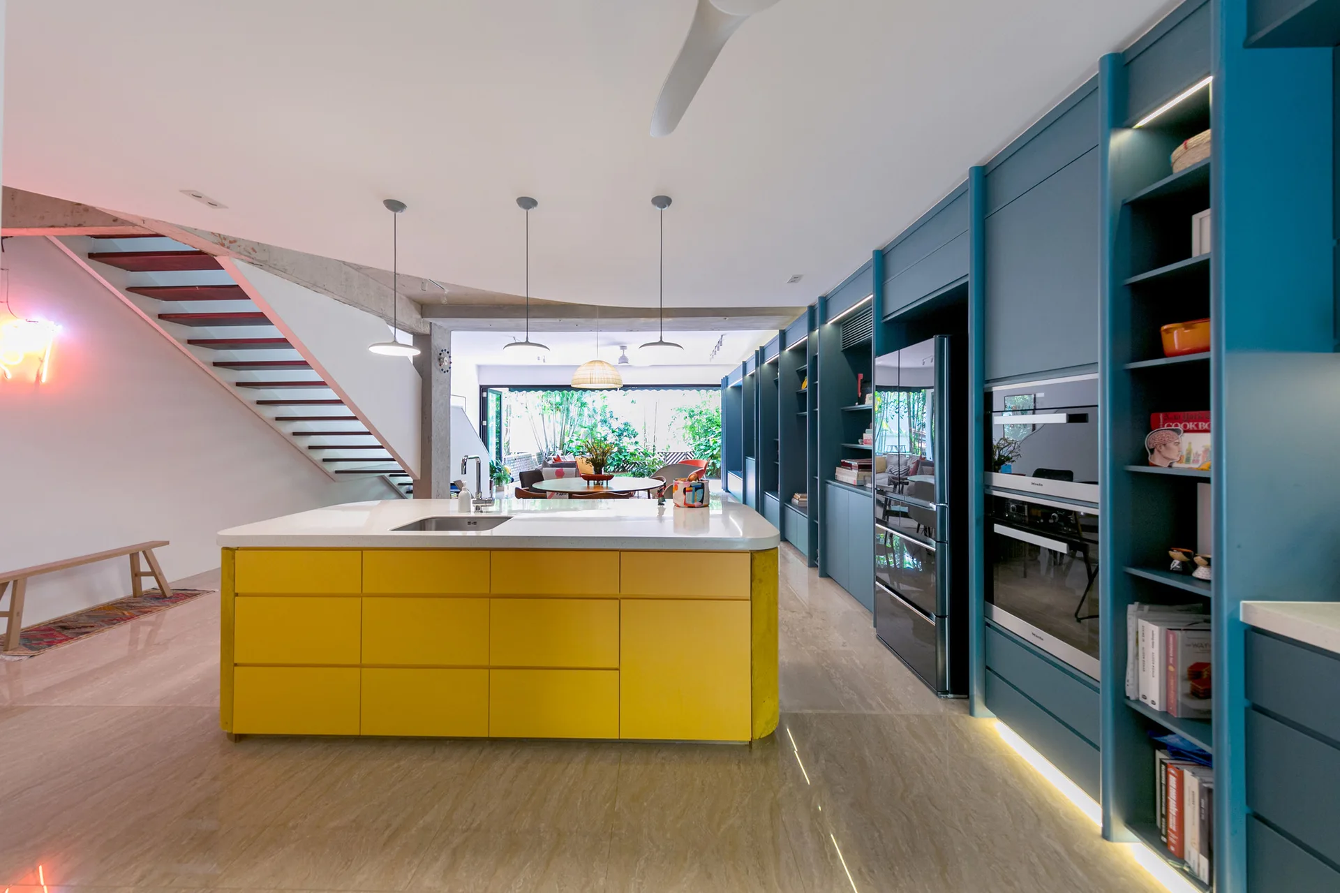 A modern open kitchen features a bright yellow island with a white countertop and sink, set against a long wall of deep teal built-in cabinets.