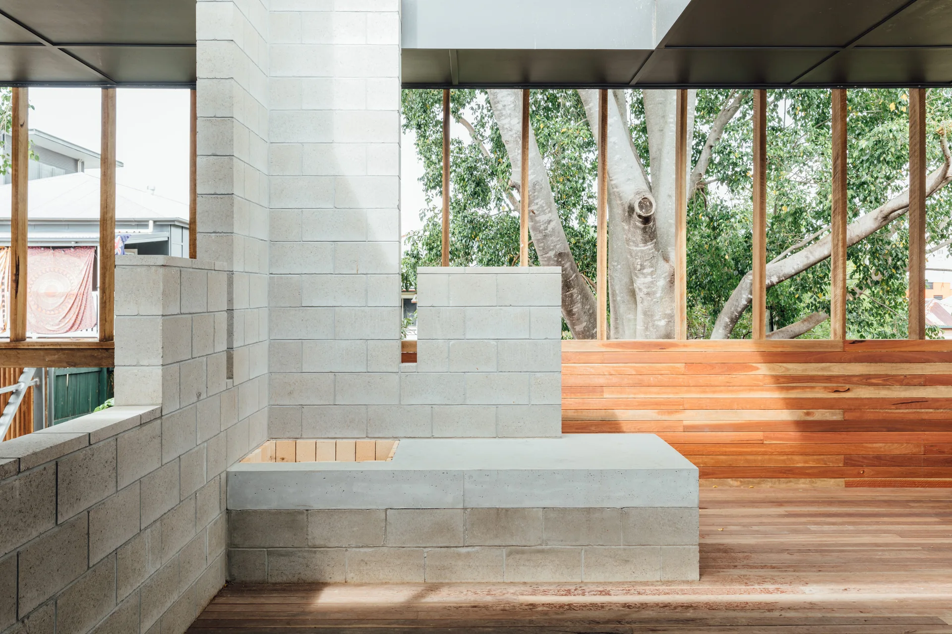 An open-air wooden terrace featuring a large concrete block fireplace structure, natural timber decking, and slatted walls framing outdoor tree views.